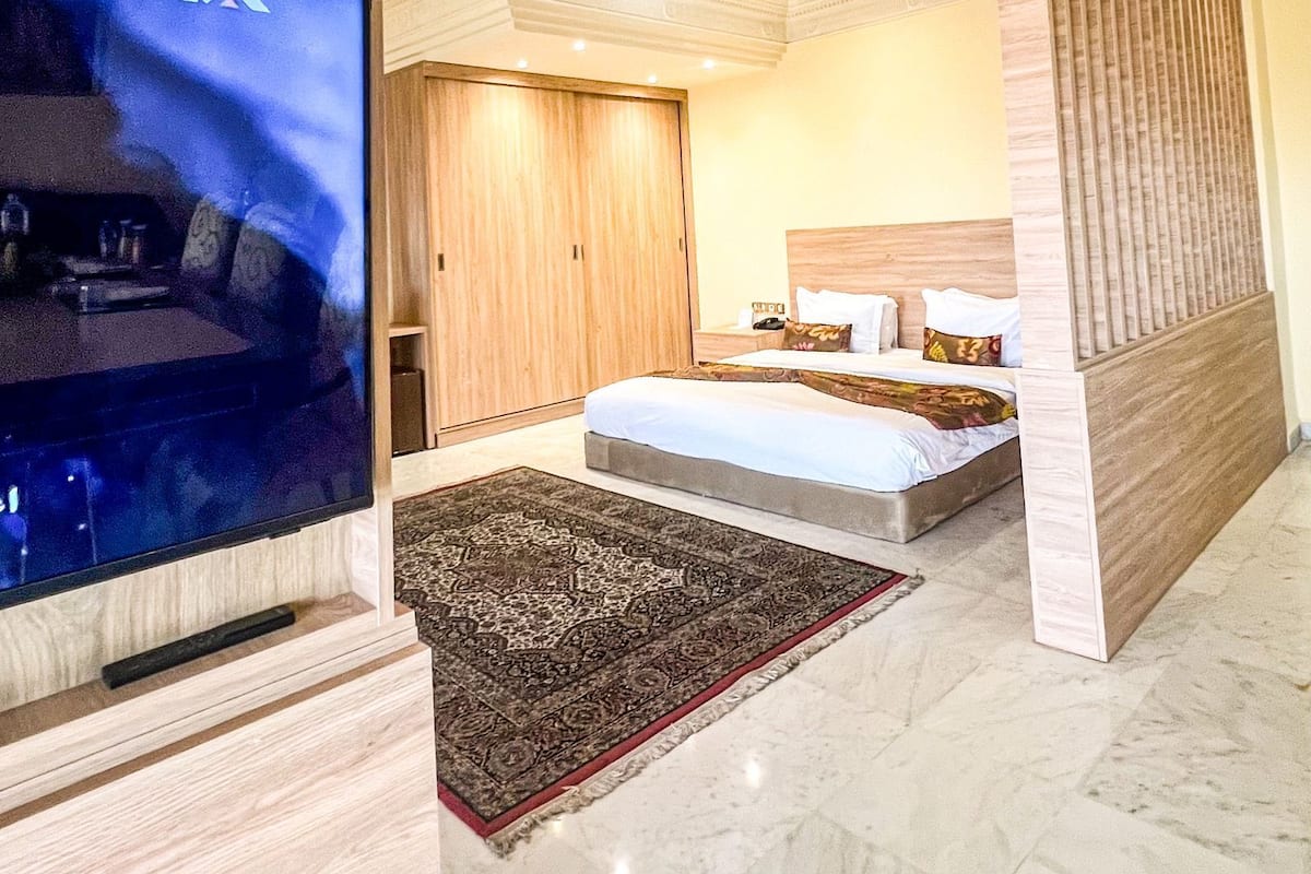 A hotel room with a large flat-screen TV, a bed with a headboard, a wooden wardrobe, and a patterned rug.