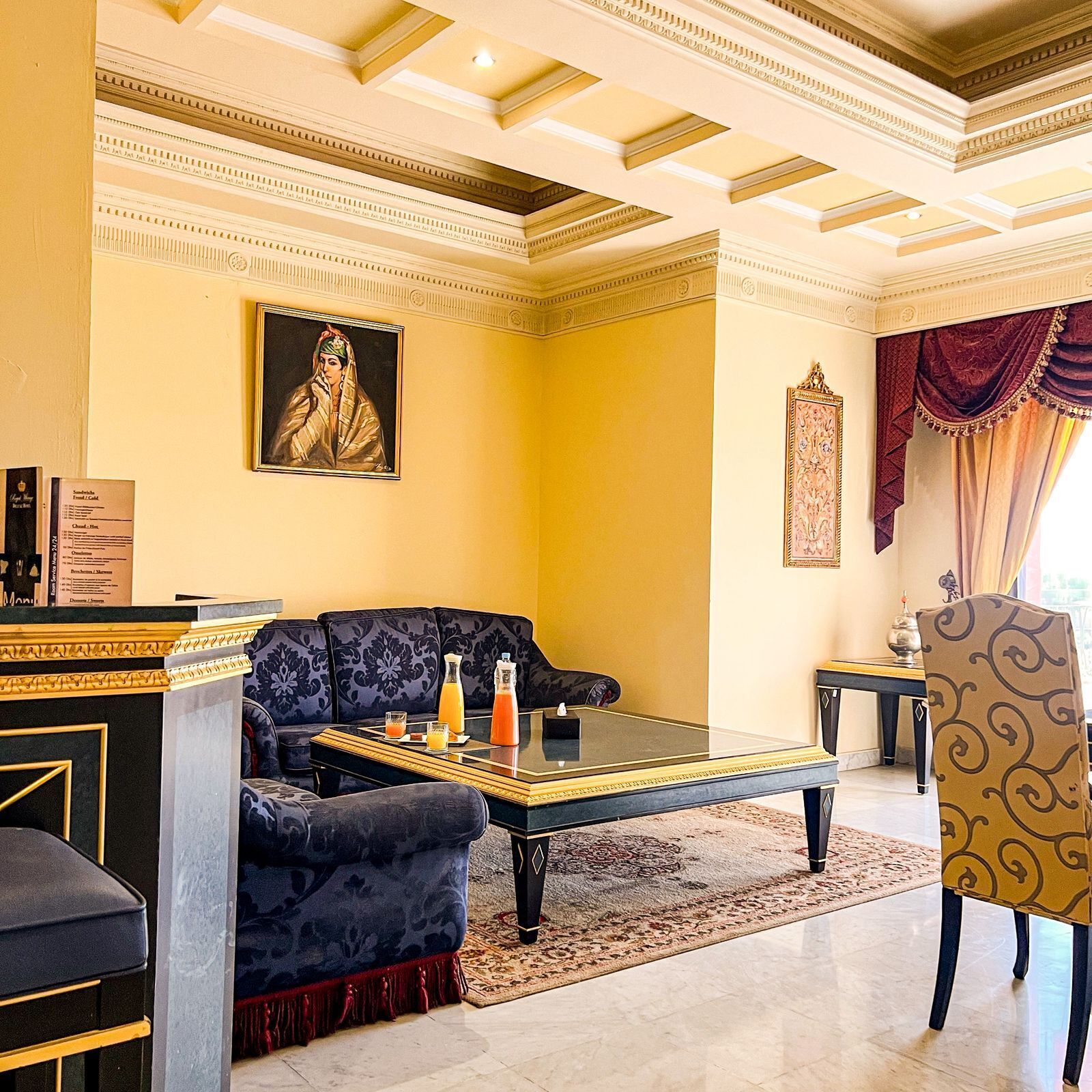 Una habitación lujosa con un sofá azul oscuro, una mesa de centro de cristal y un sillón estampado. Se observa un cuadro en la pared y un espejo decorativo.