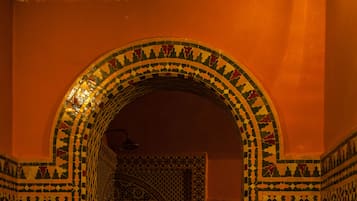 A narrow hallway with intricate geometric tile patterns on the walls and floor, leading to a room with a patterned ceiling.
