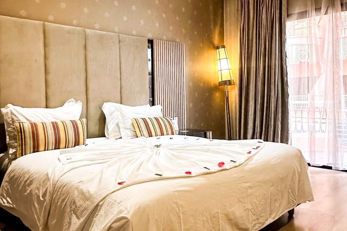 A hotel room with a large bed, two pillows, a bedside lamp, and a view of the outside through a window with curtains.