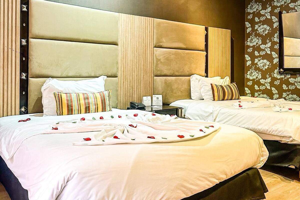 A hotel room with two beds, each with a large padded headboard, white bedding, and a floral wallpaper design.