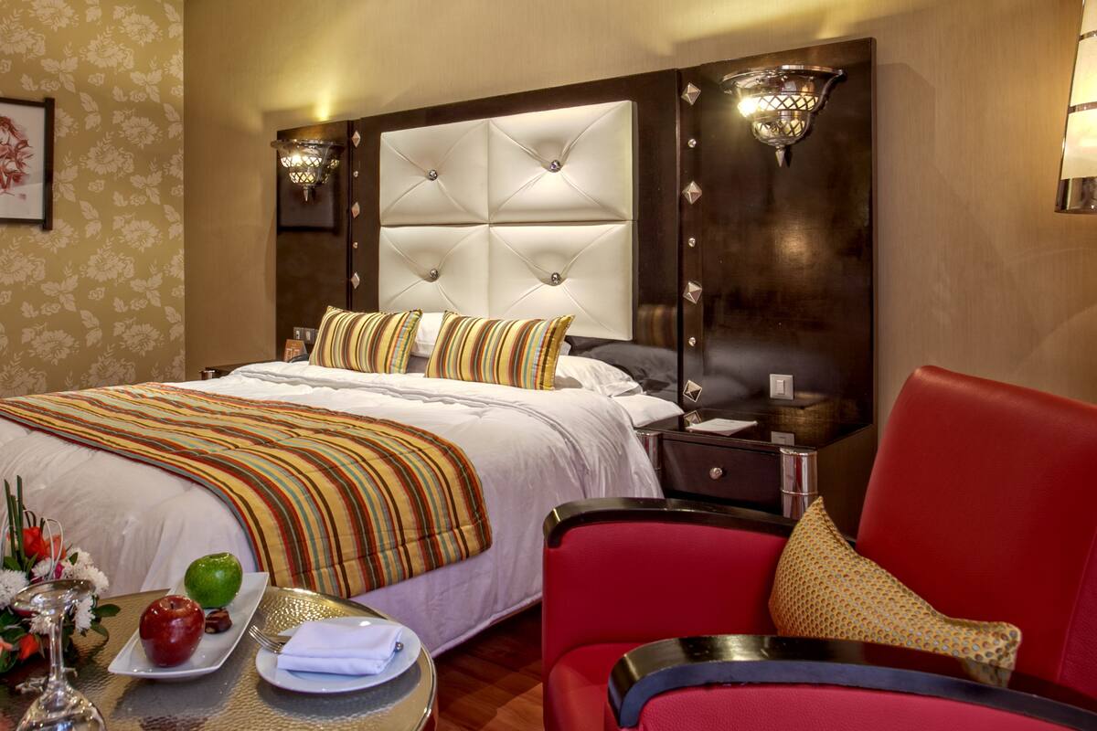 A hotel room with a large bed, a red armchair, a bedside table with a lamp, and a floral wallpaper design.