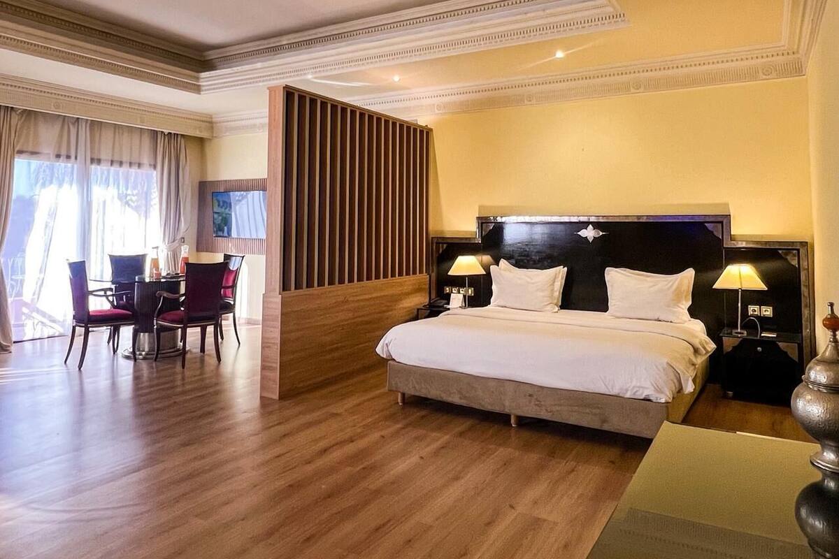 A hotel room with a large bed, a desk with chairs, a chandelier, and a view of the outdoors.