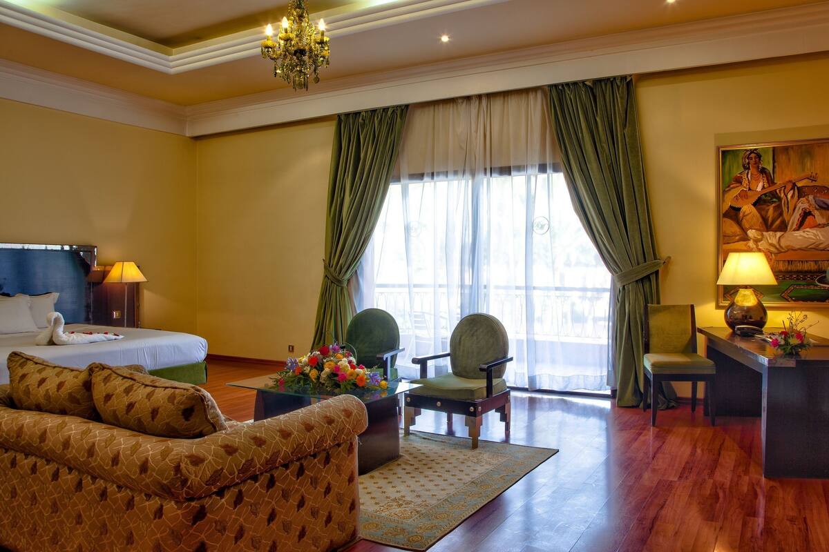 A hotel room with a bed, a sofa, a desk with a lamp, a painting, and a window with curtains.