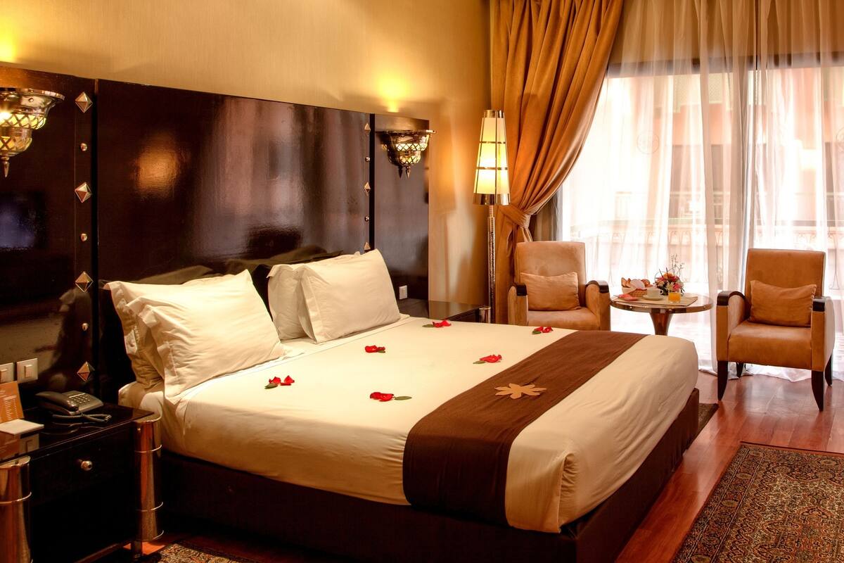 A hotel room with a large bed, two armchairs, a small table, a TV, and a view of the city through sheer curtains.