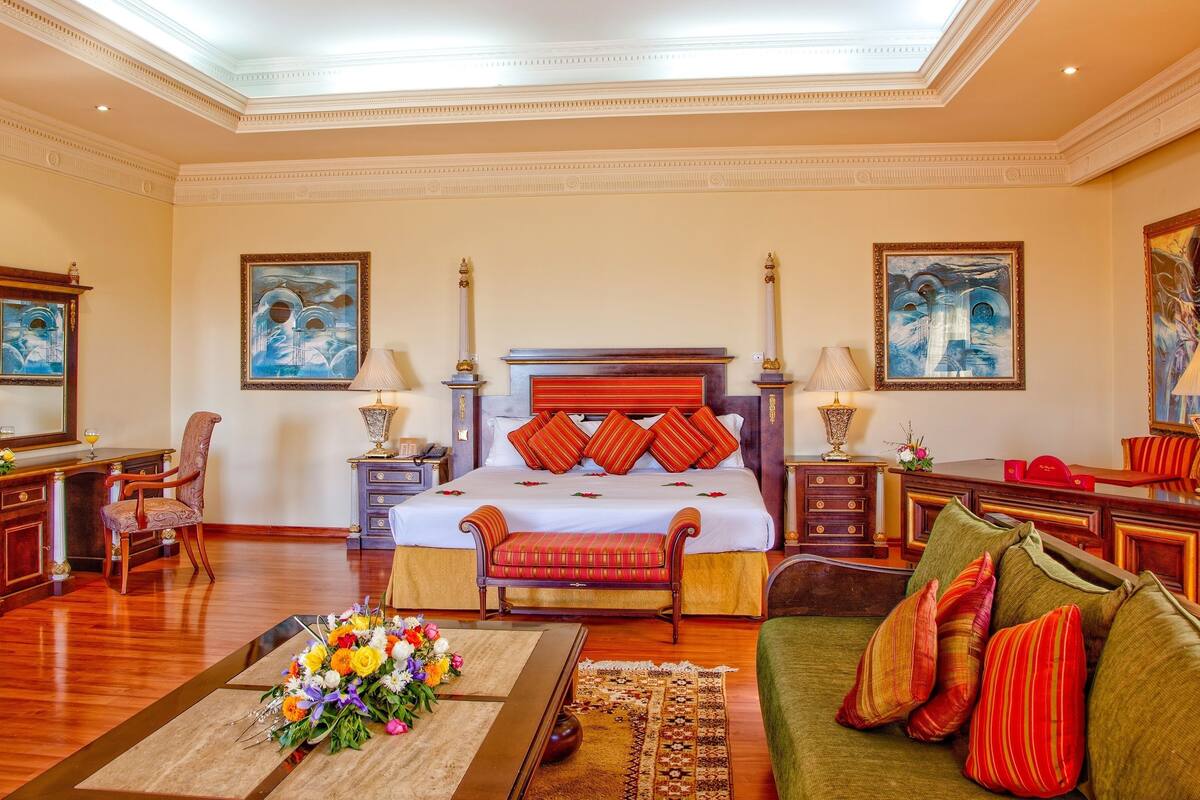 A spacious hotel room with a large bed, a sitting area with a sofa and armchairs, a desk, and a dining table.