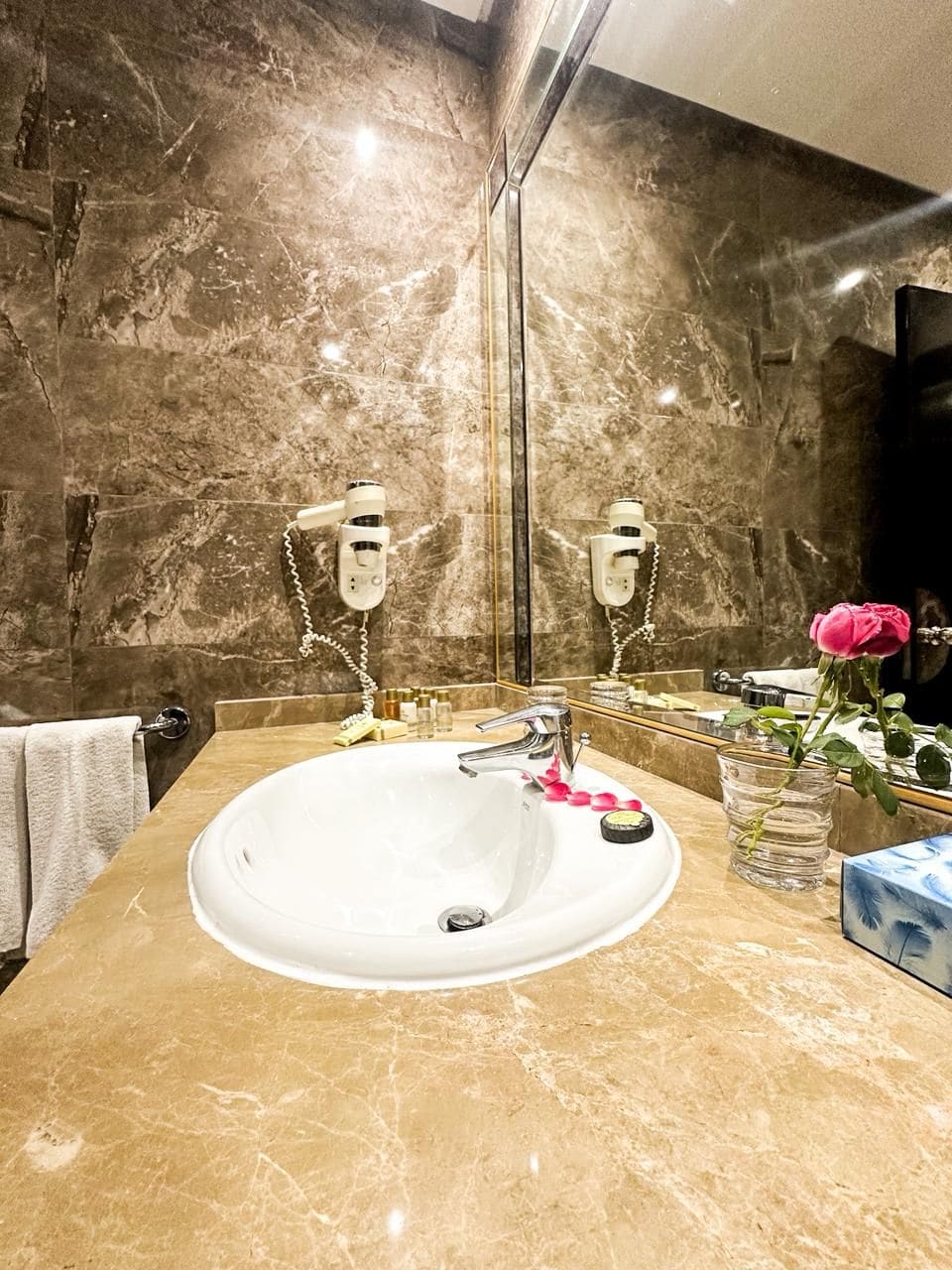 Une salle de bain avec un lavabo en marbre, deux appliques murales, un miroir et un vase contenant une rose rose.