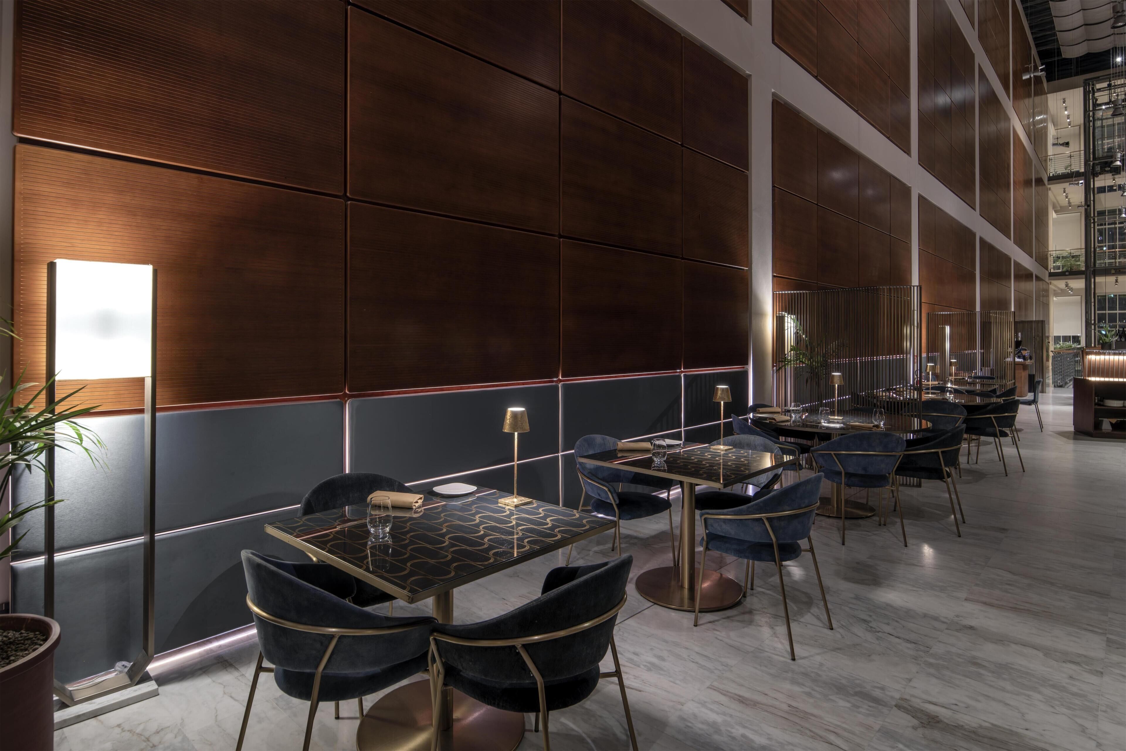 A modern interior with a long dining area featuring tables and chairs, a large window, and a high ceiling.