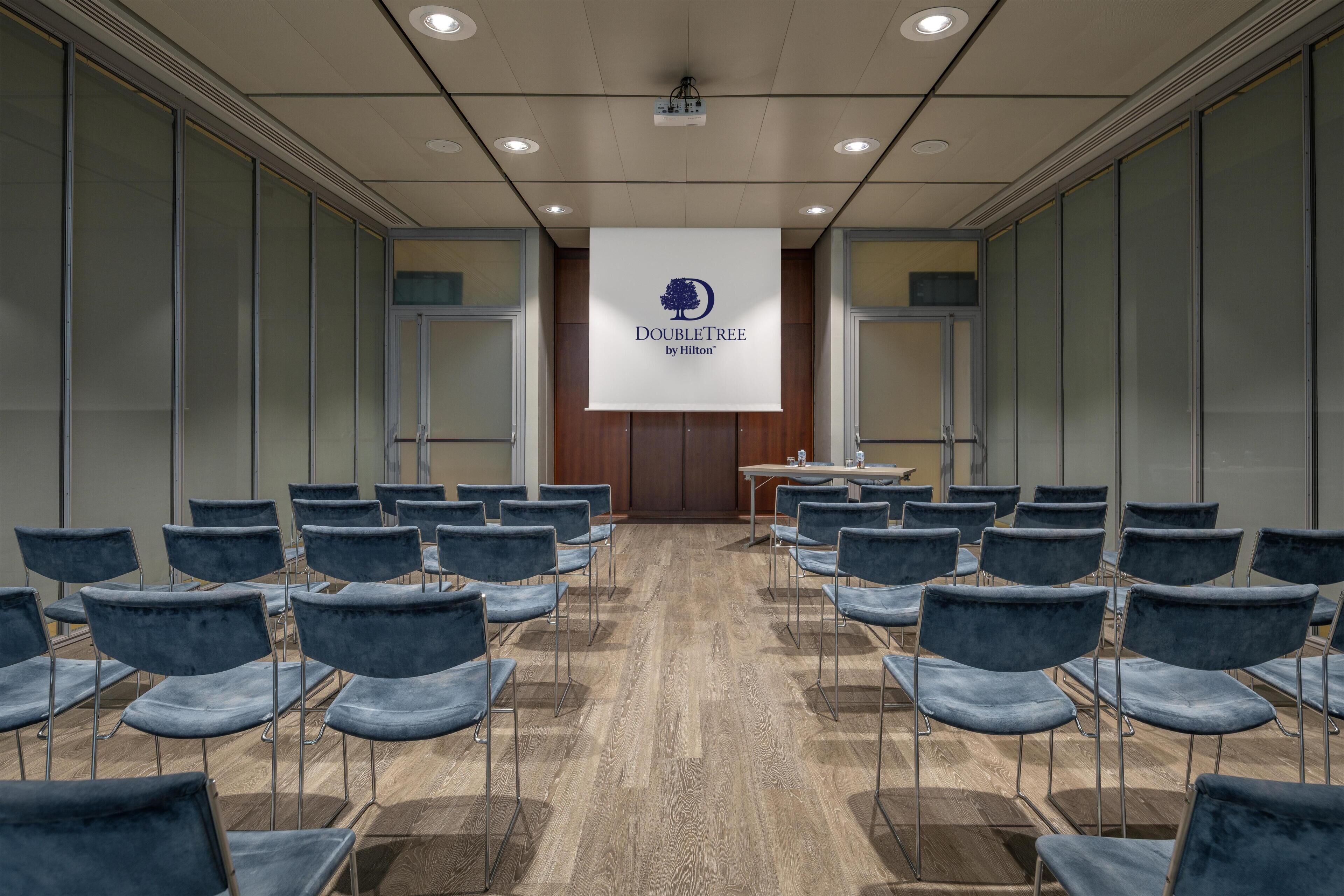 A conference room with a presentation screen displaying the DoubleTree by Hilton logo, rows of blue chairs, and a wooden floor.