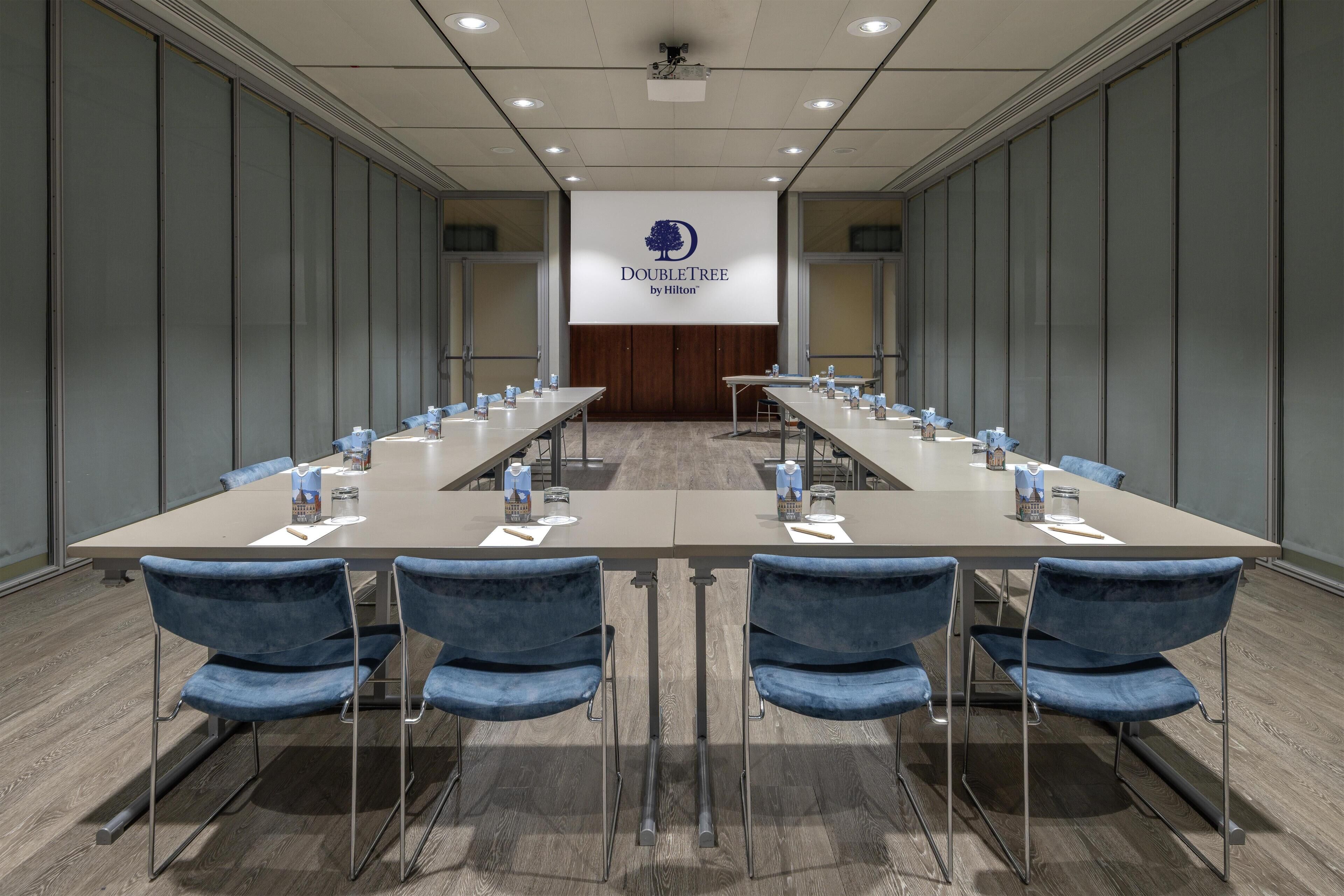 Una sala conferenze con un lungo tavolo, sedie blu e uno schermo proiettore che mostra 'DOUBLETREE by Hilton'.