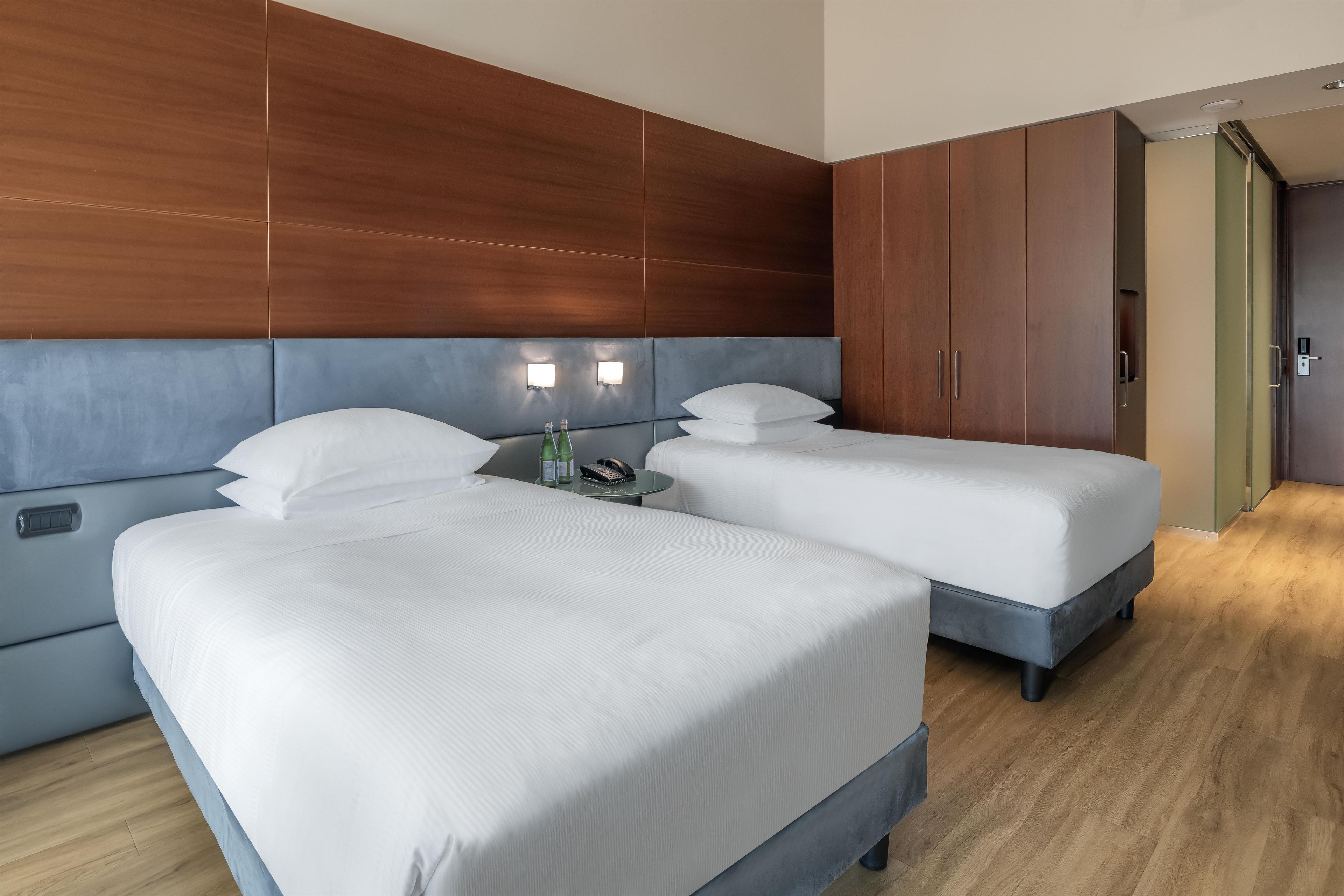 A hotel room with two beds, a wooden headboard, a nightstand with a phone and bottles, and a door leading to another room.