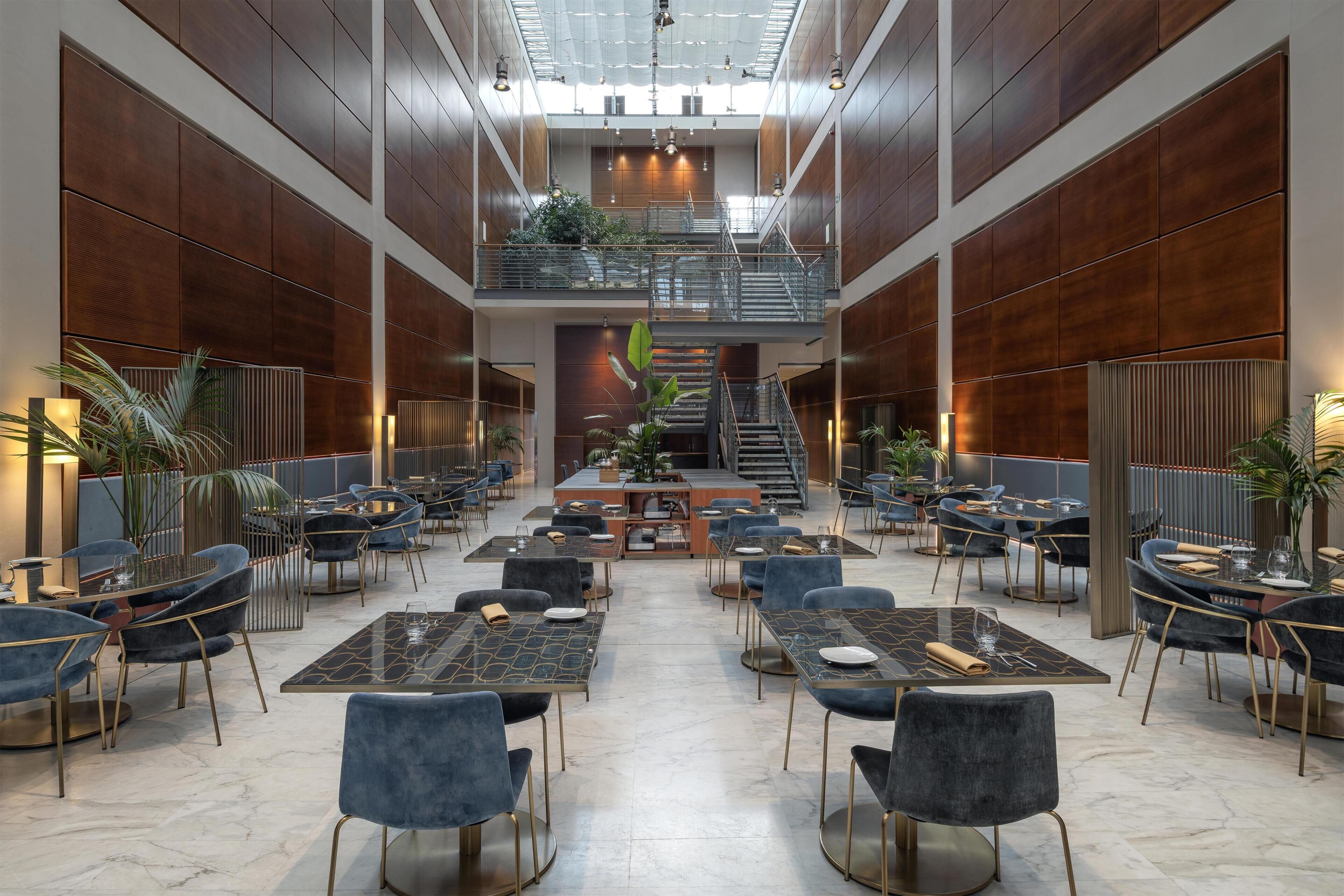 A spacious indoor area with multiple dining tables and chairs, a central staircase, and a mezzanine level with a railing.