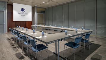 Un salón de conferencias con una larga mesa, sillas, una pantalla de proyector y un ventanal.