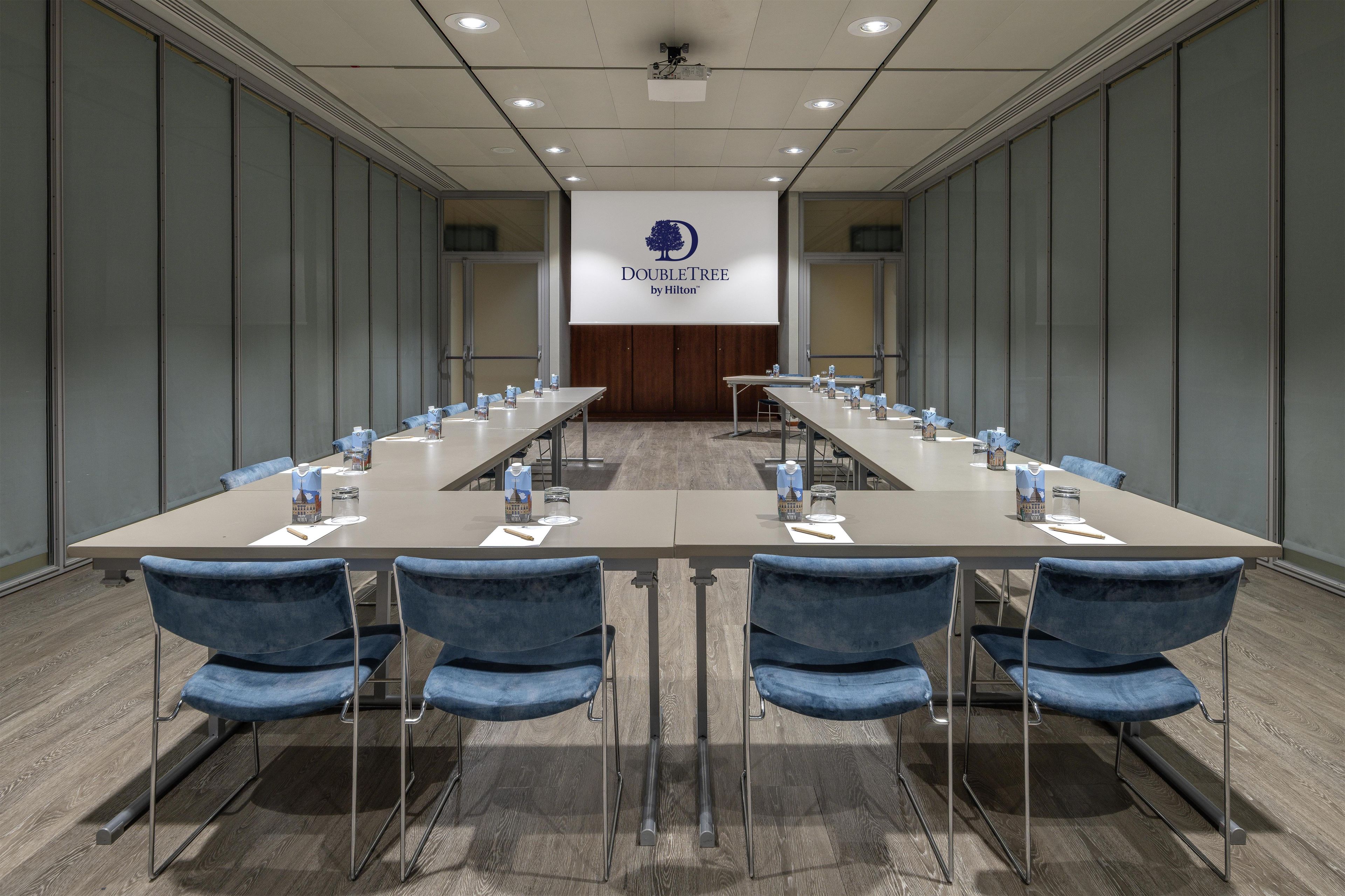 Une salle de conférence avec une longue table, des chaises bleues et un écran de projection affichant le logo « DOUBLETREE by Hilton ».