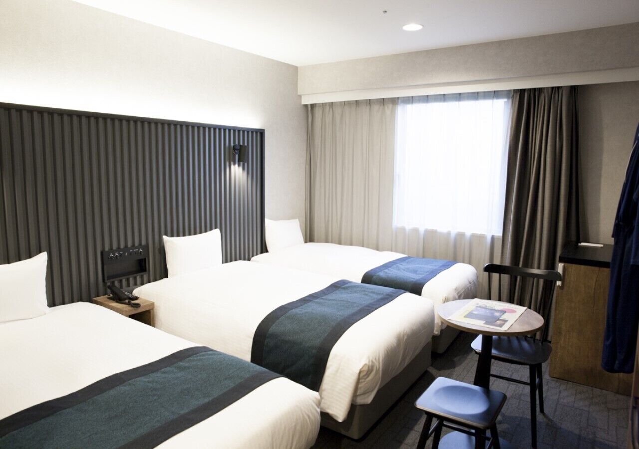 A hotel room with two beds, a small table, a chair, a desk, and a window with curtains.