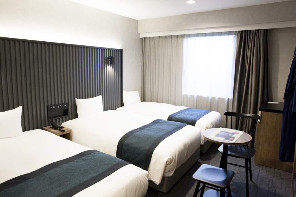 A hotel room with two beds, a small table, a chair, a desk, and a window with curtains.