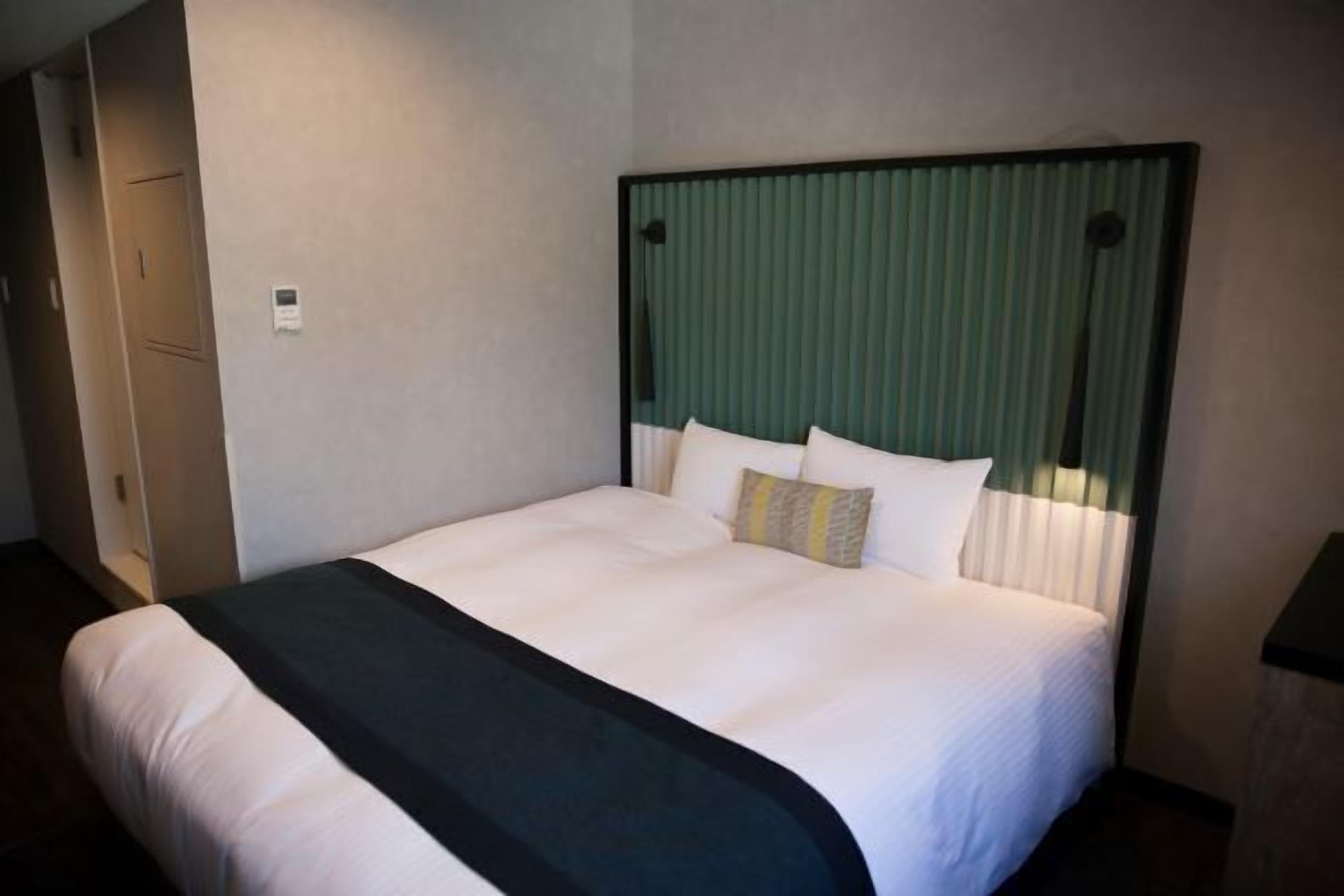 A hotel room with a large bed, a headboard with vertical blinds, a bedside table, and a wardrobe.