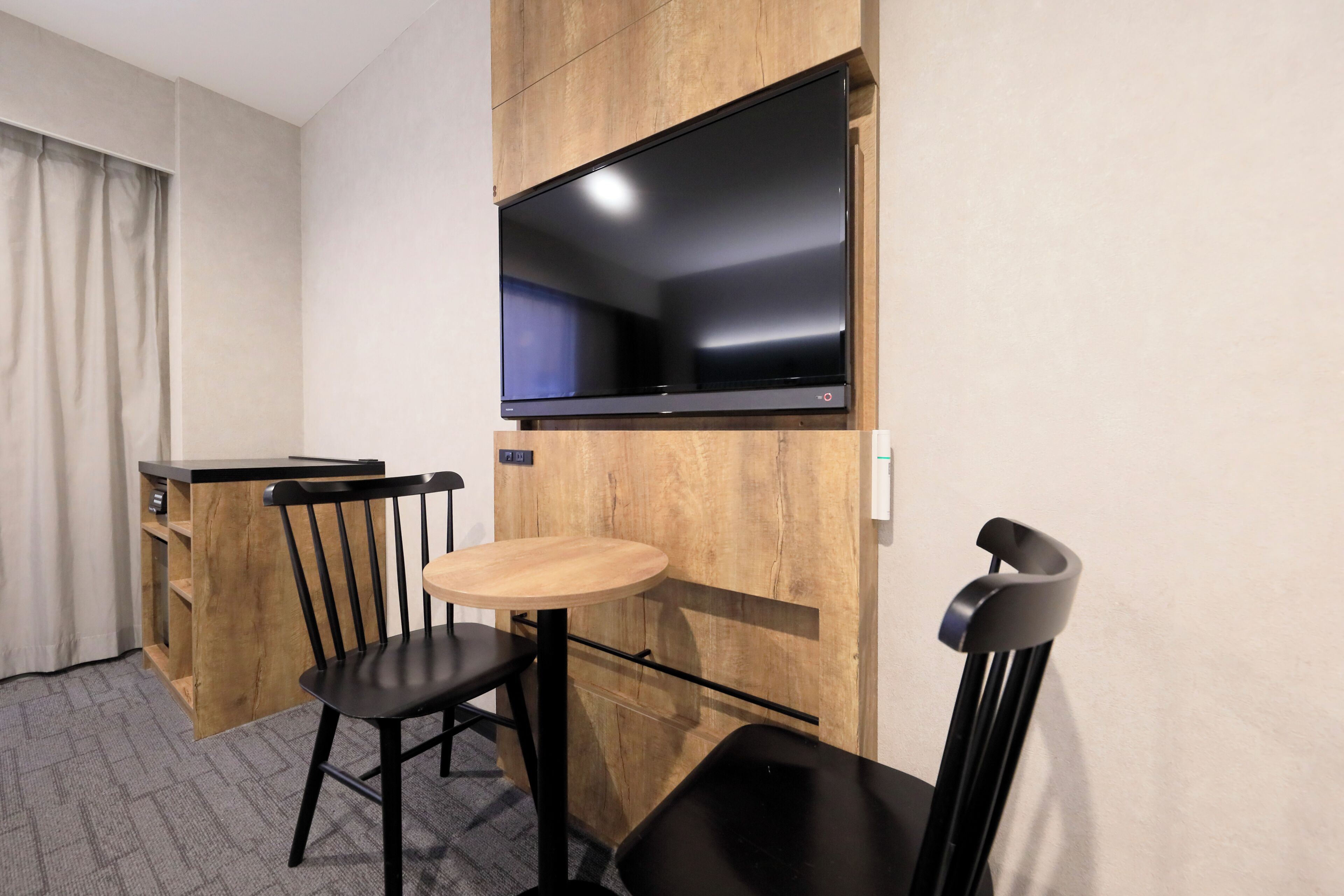 A compact hotel room with a kitchenette, a dining area with a table and chairs, and a large flat-screen TV mounted above the counter.