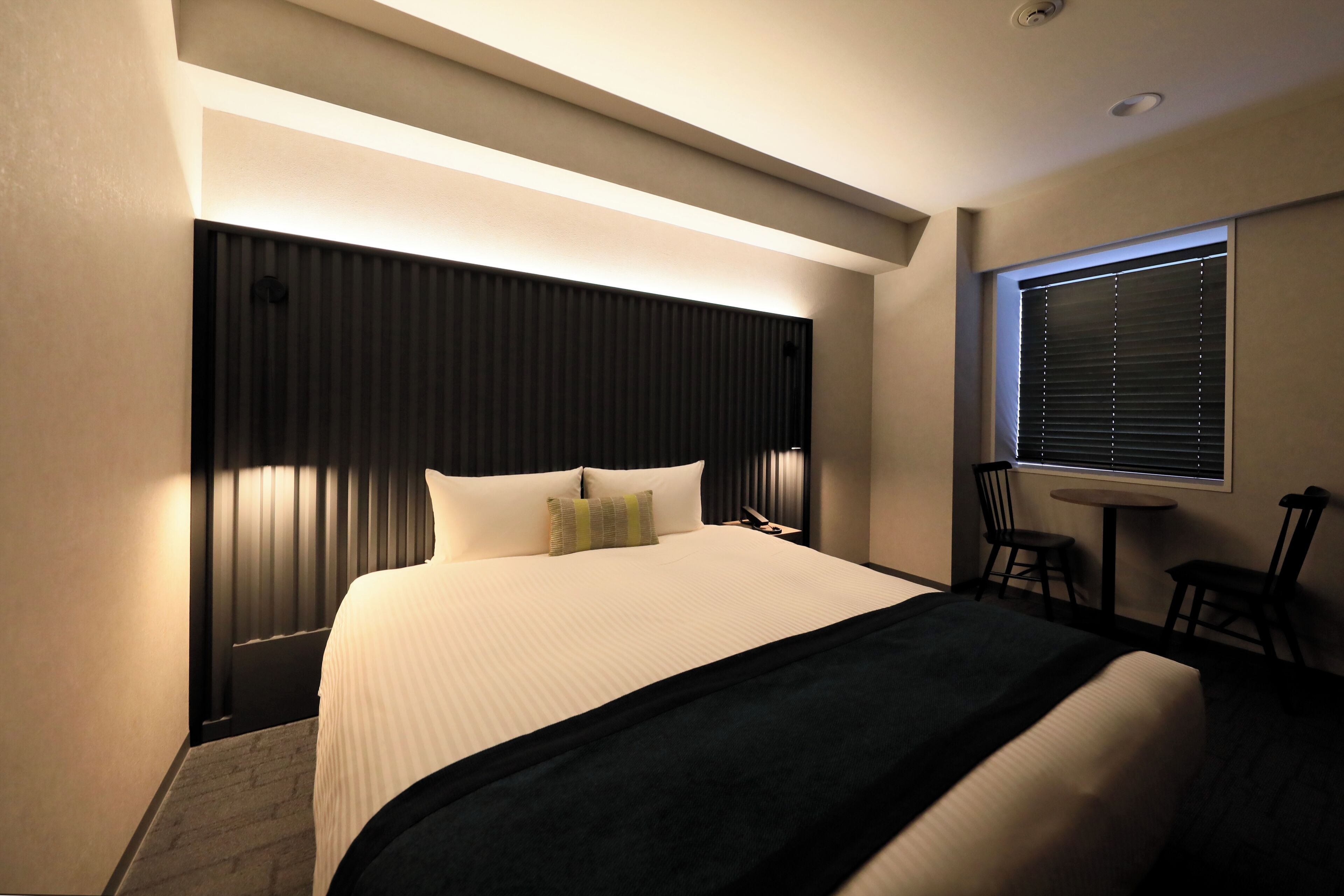 A hotel room with a large bed, a bedside table, a chair, and a window with blinds.