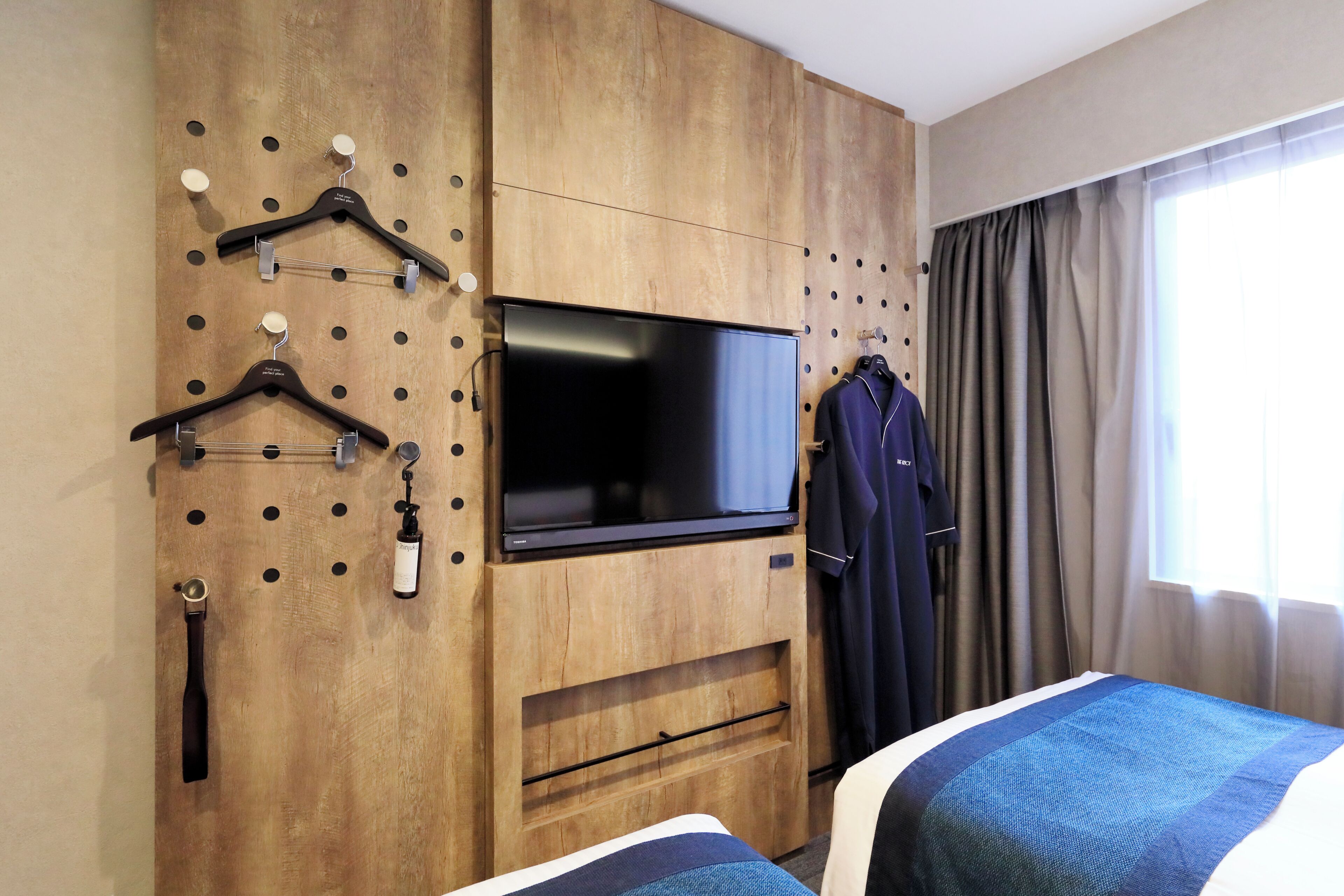 A hotel room with a wooden wardrobe, a flat-screen TV, a coat rack, and a bed with a blue and white bedspread.