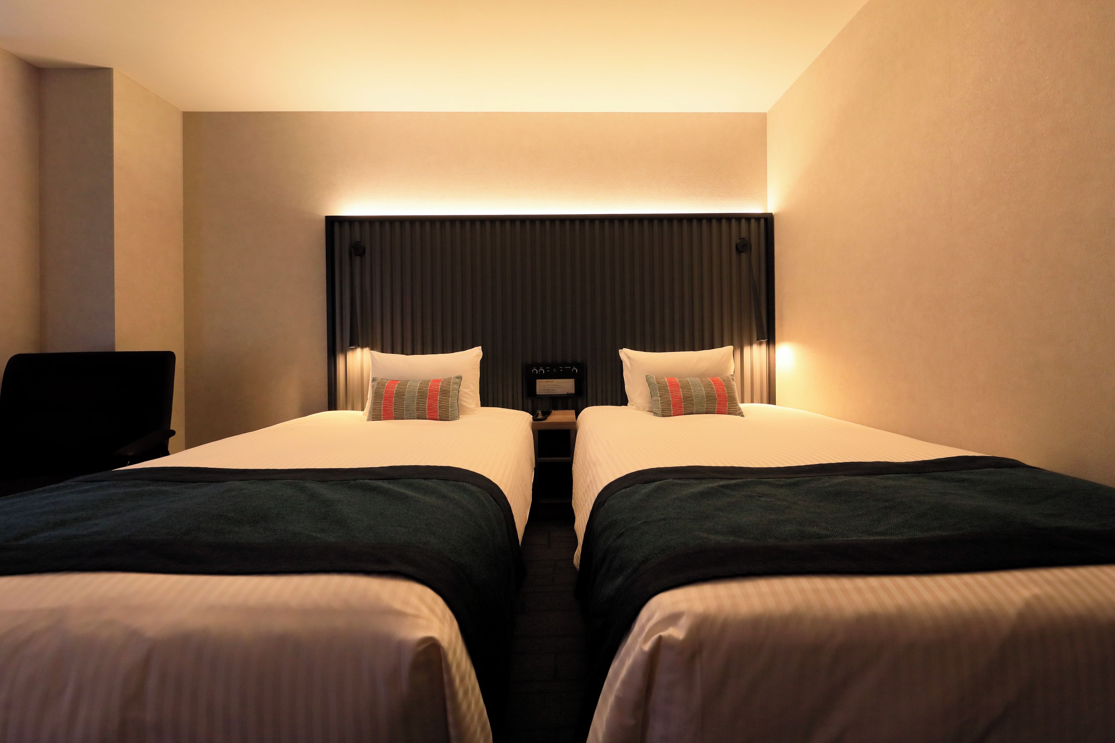 A hotel room with two beds, a desk, and a wall-mounted control panel.