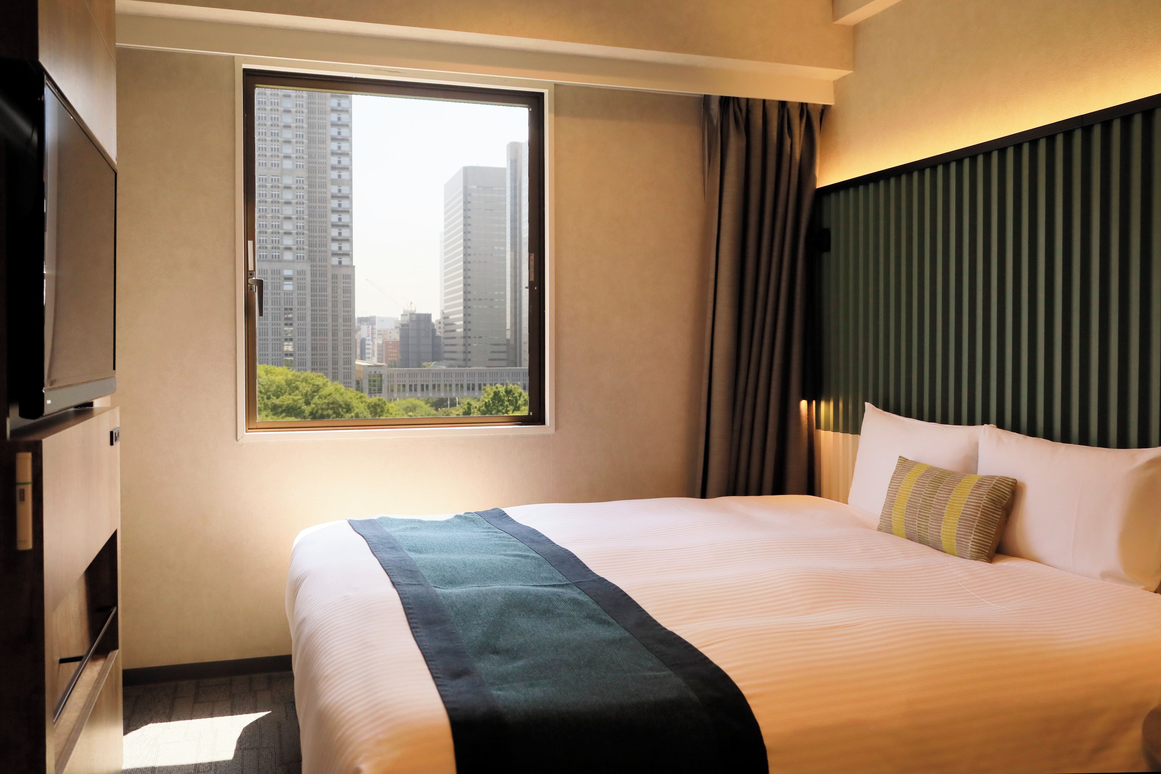 A hotel room with a bed, a TV, a window with a city view, and a curtain.