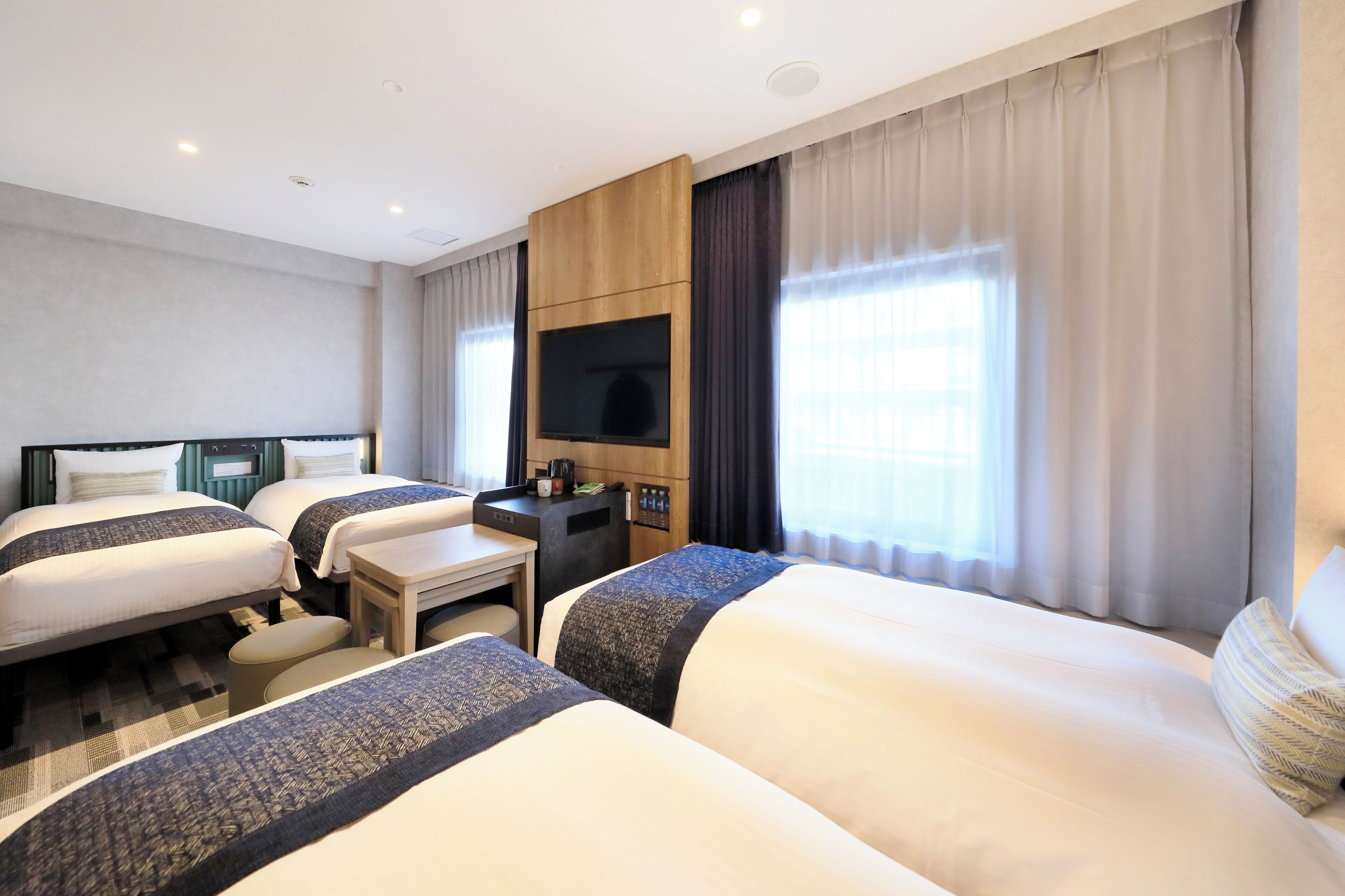 A hotel room with two beds, a desk, a flat-screen TV, and a large window with curtains.