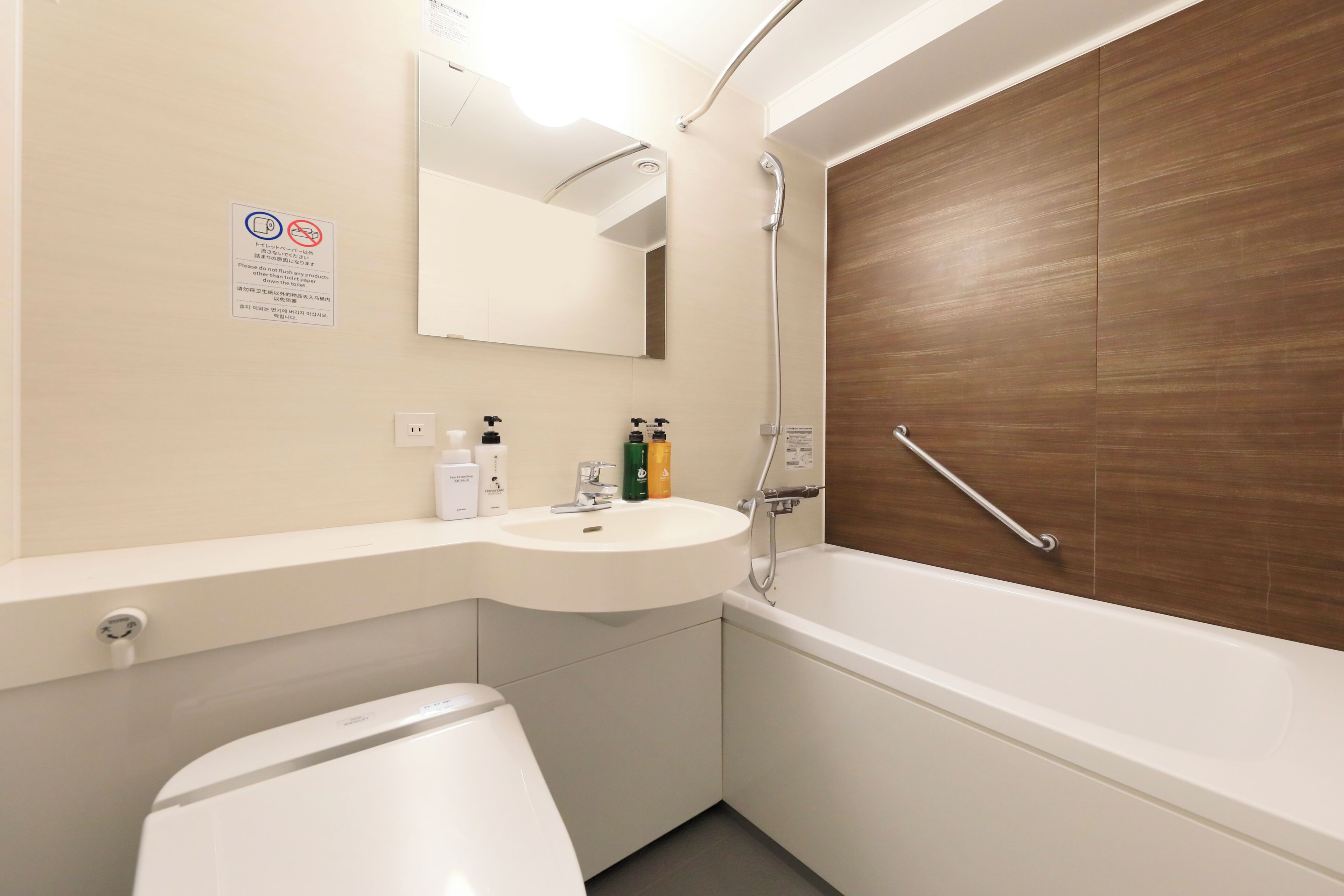 Un baño moderno con lavamanos, espejo y retrete blancos. Se observa una puerta de madera y una zona de ducha con barra de apoyo.