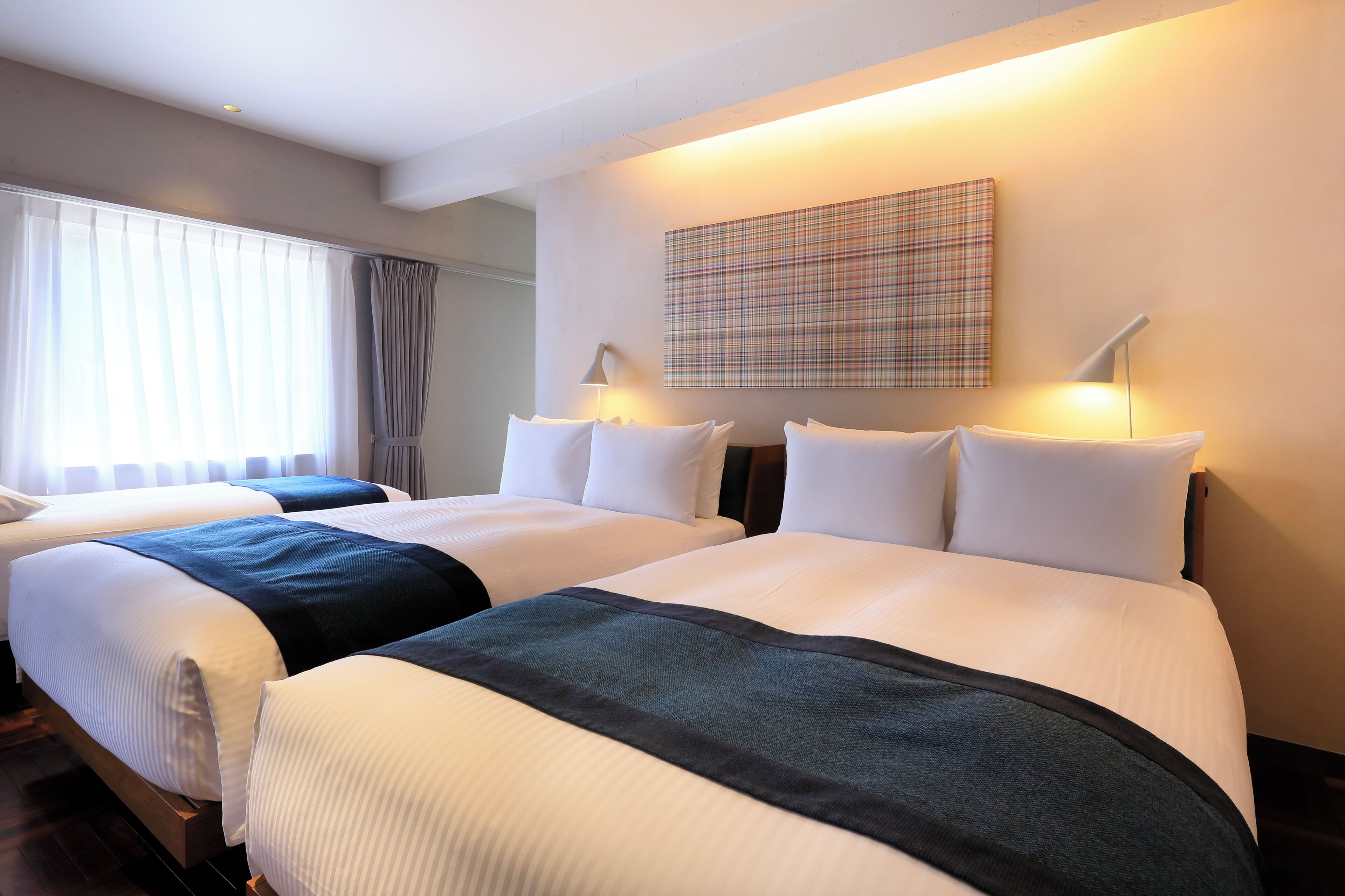 Habitación de hotel con dos camas, un ventanal con cortinas, una obra de arte colgada en la pared y una mesita de noche con lámpara.