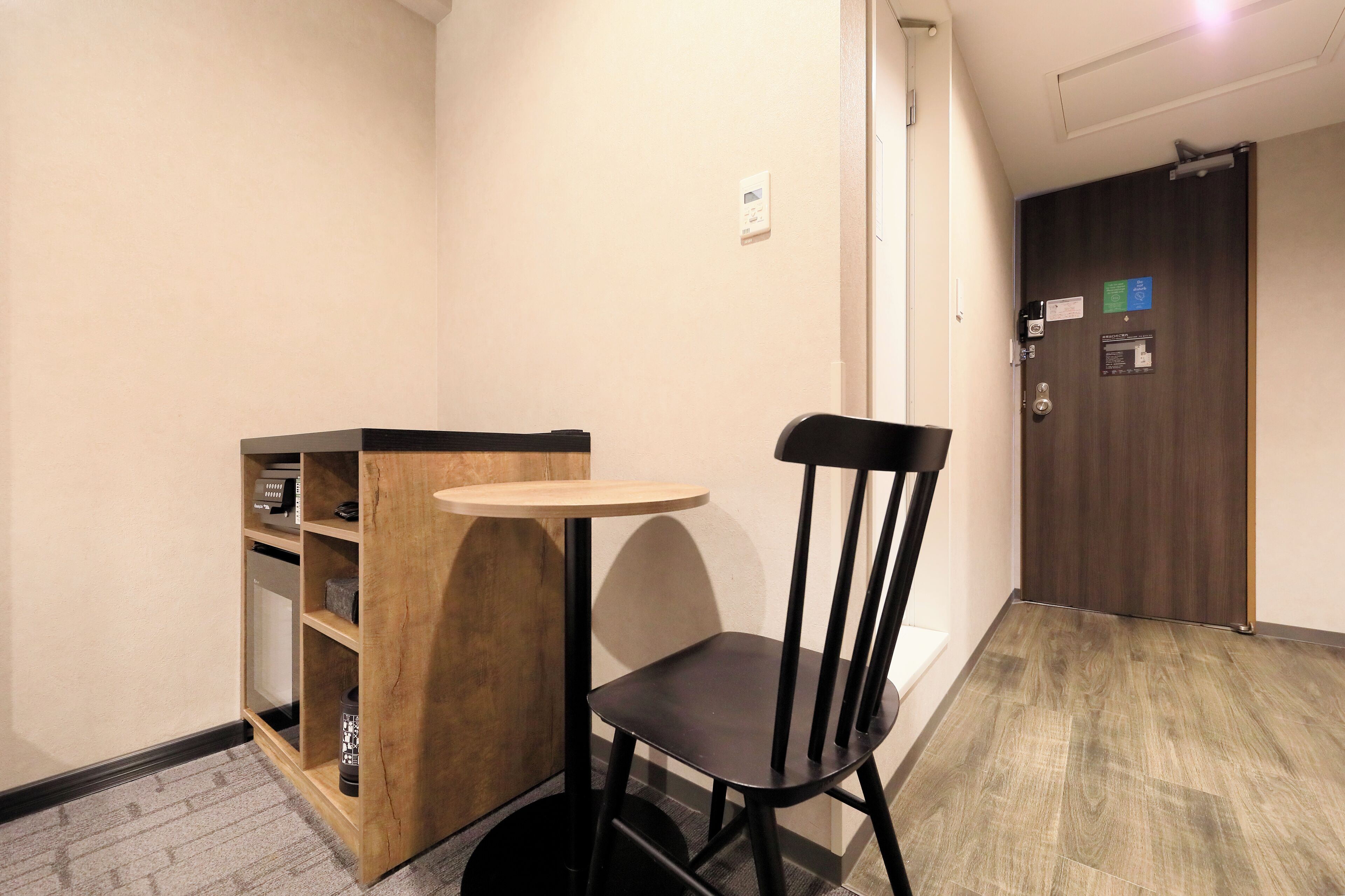 A small room with a wooden table and a black chair, a wooden cabinet with shelves, a door with a lock, and a light switch on the wall.