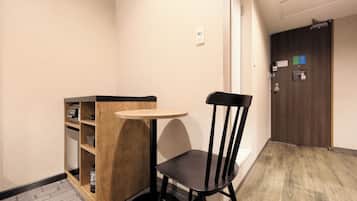 Una habitación pequeña con una mesa de madera y una silla negra, un armario de madera con estantes, una puerta con cerradura y un interruptor de luz en la pared.