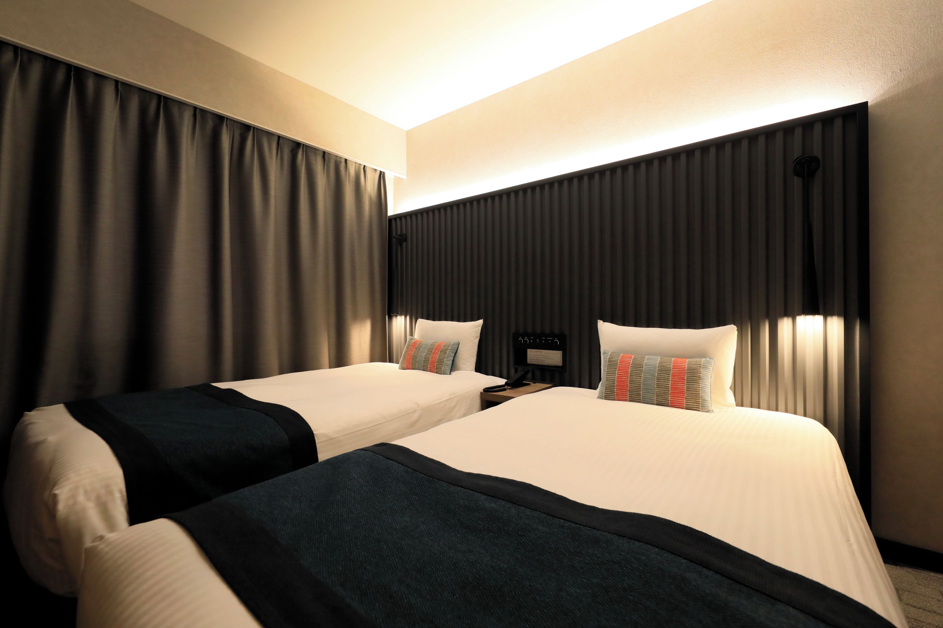 A hotel room with two beds, a nightstand with a phone, and a headboard with built-in lighting.