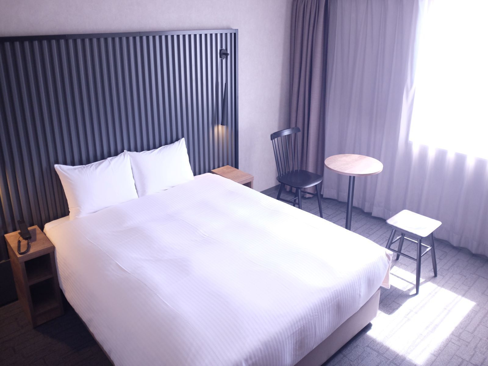 A hotel room with a large bed, a bedside table, a chair, a small round table, and a window with curtains.