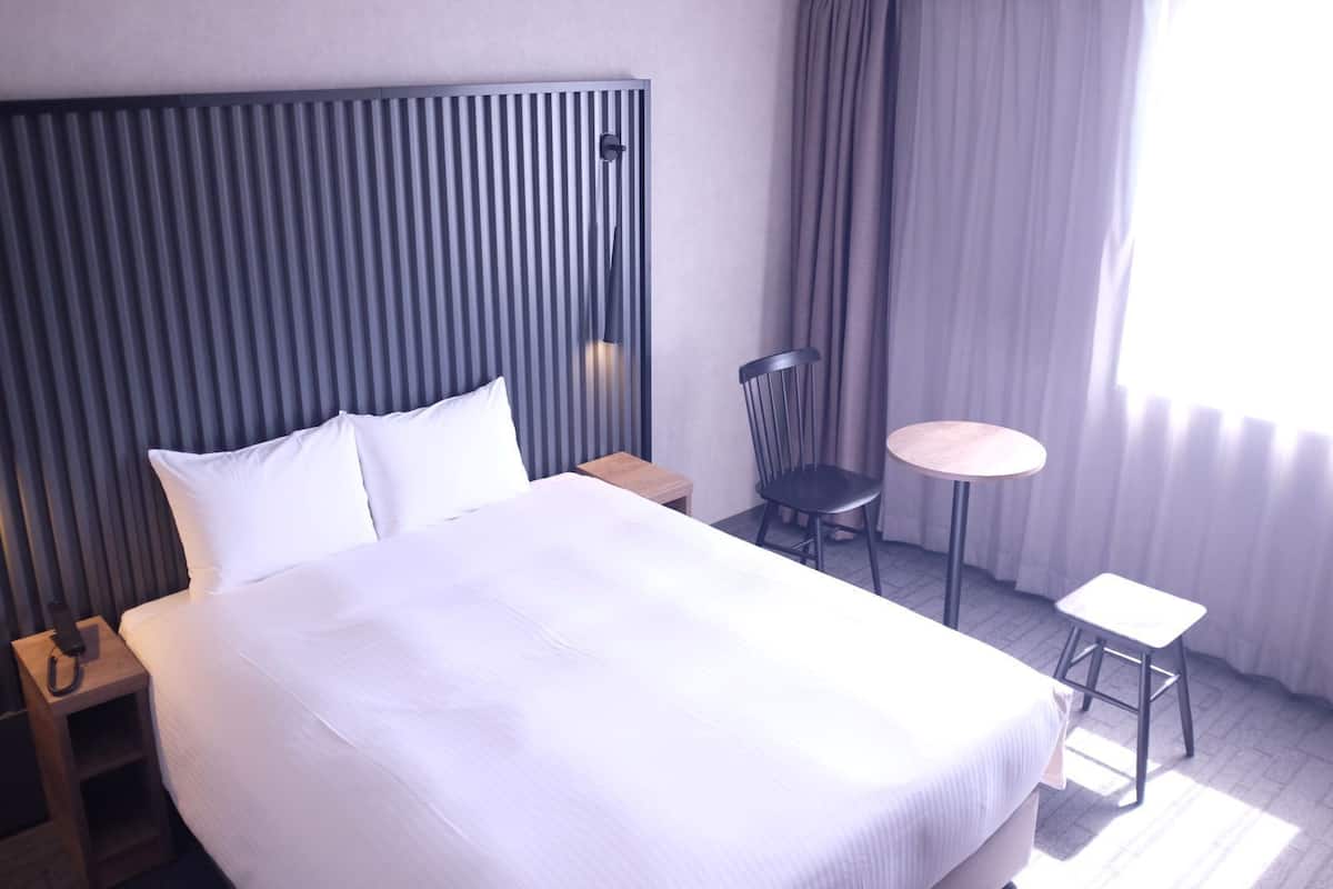 A hotel room with a large bed, a bedside table, a chair, a small round table, and a window with curtains.