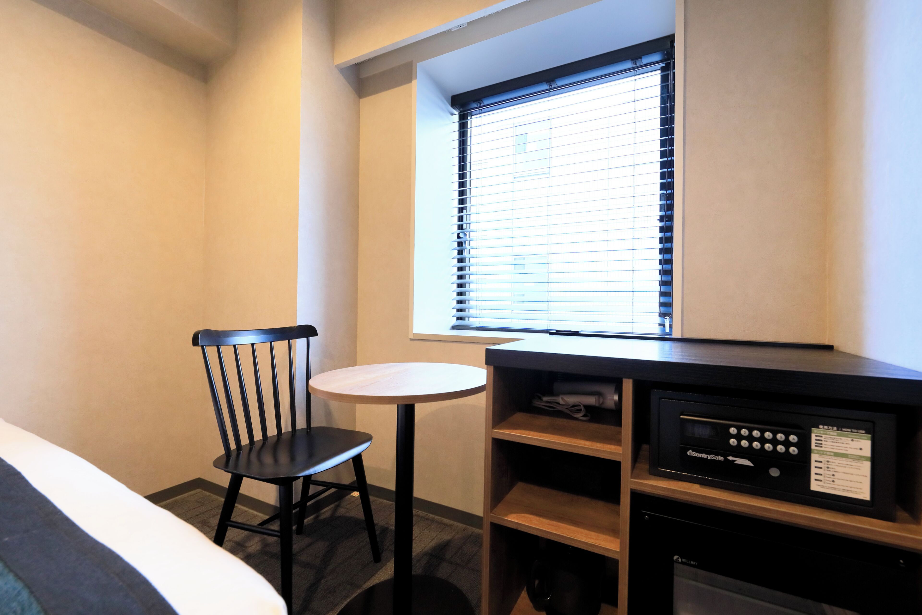 Ein kleines Zimmer mit Bett, Stuhl, Tisch, einem Fenster mit Jalousien und einer Minibar.