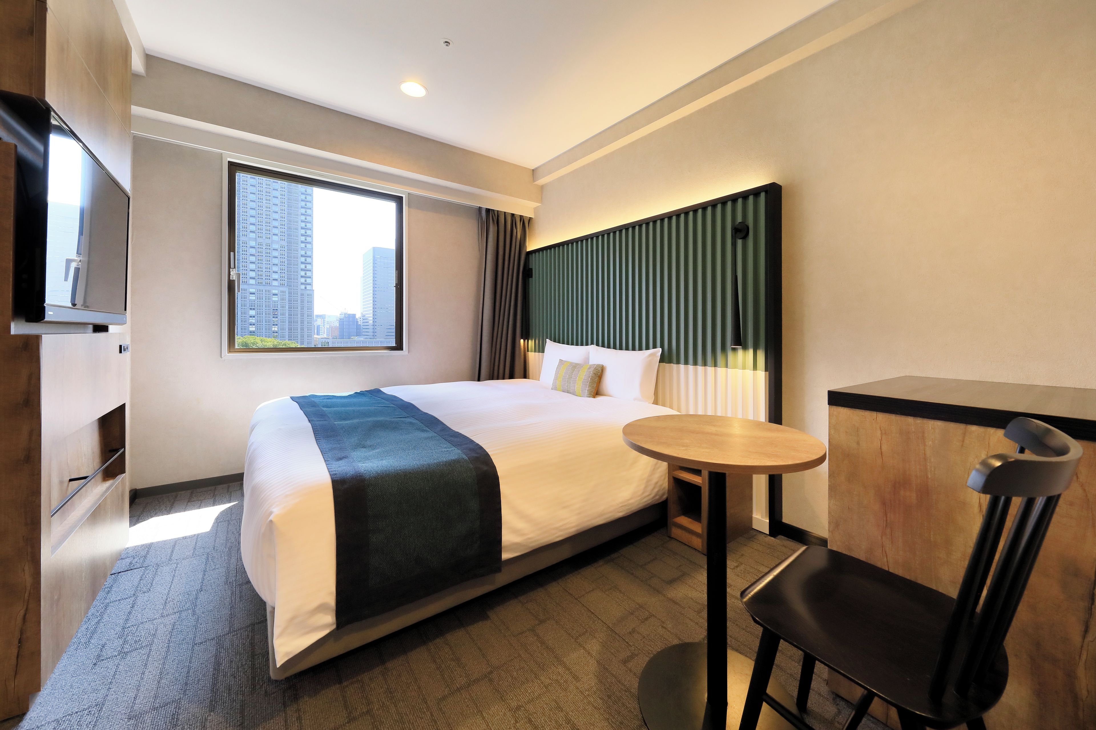 A hotel room with a bed, a small table, a chair, a TV, and a window with a city view.