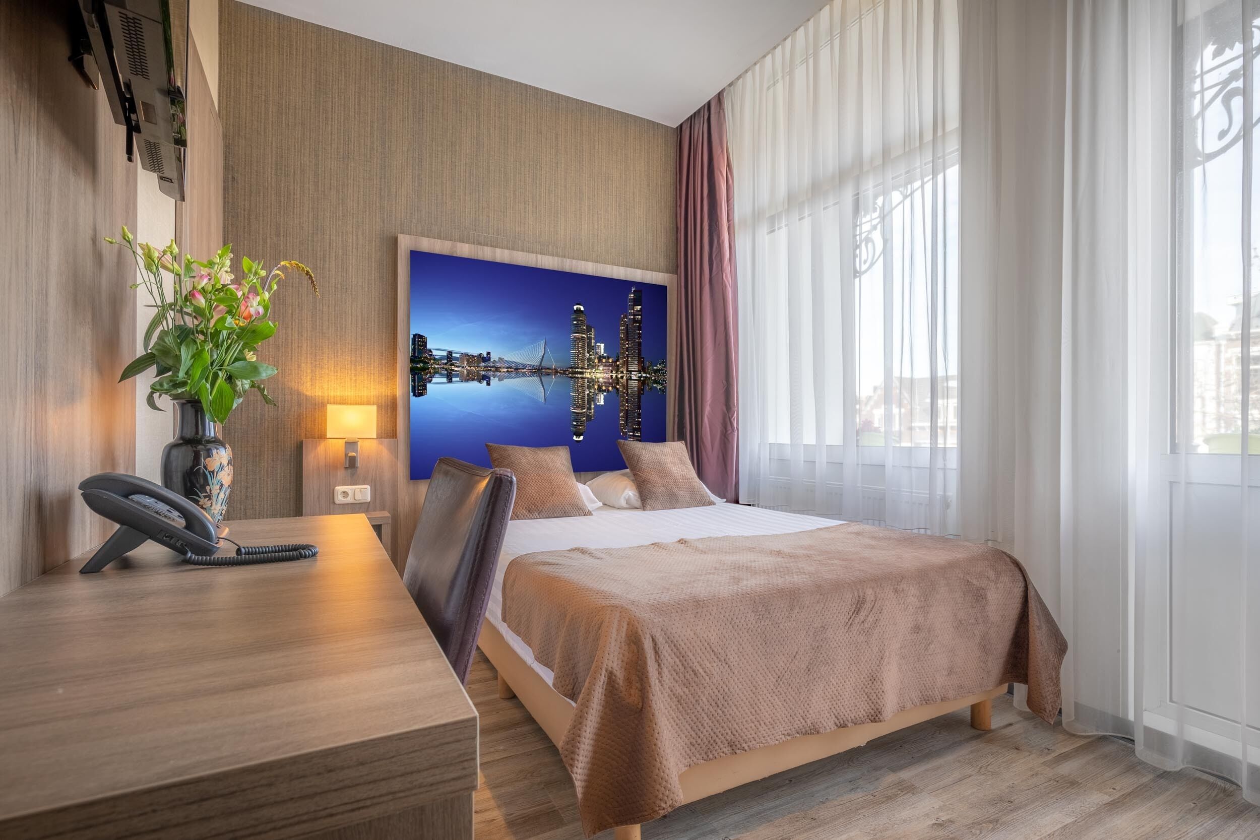 A hotel room with a bed, a desk with a telephone, a vase with flowers, a wall-mounted air conditioner, and a cityscape painting.