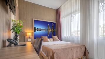 A hotel room with a bed, a desk with a telephone, a vase with flowers, a wall-mounted air conditioner, and a cityscape painting.