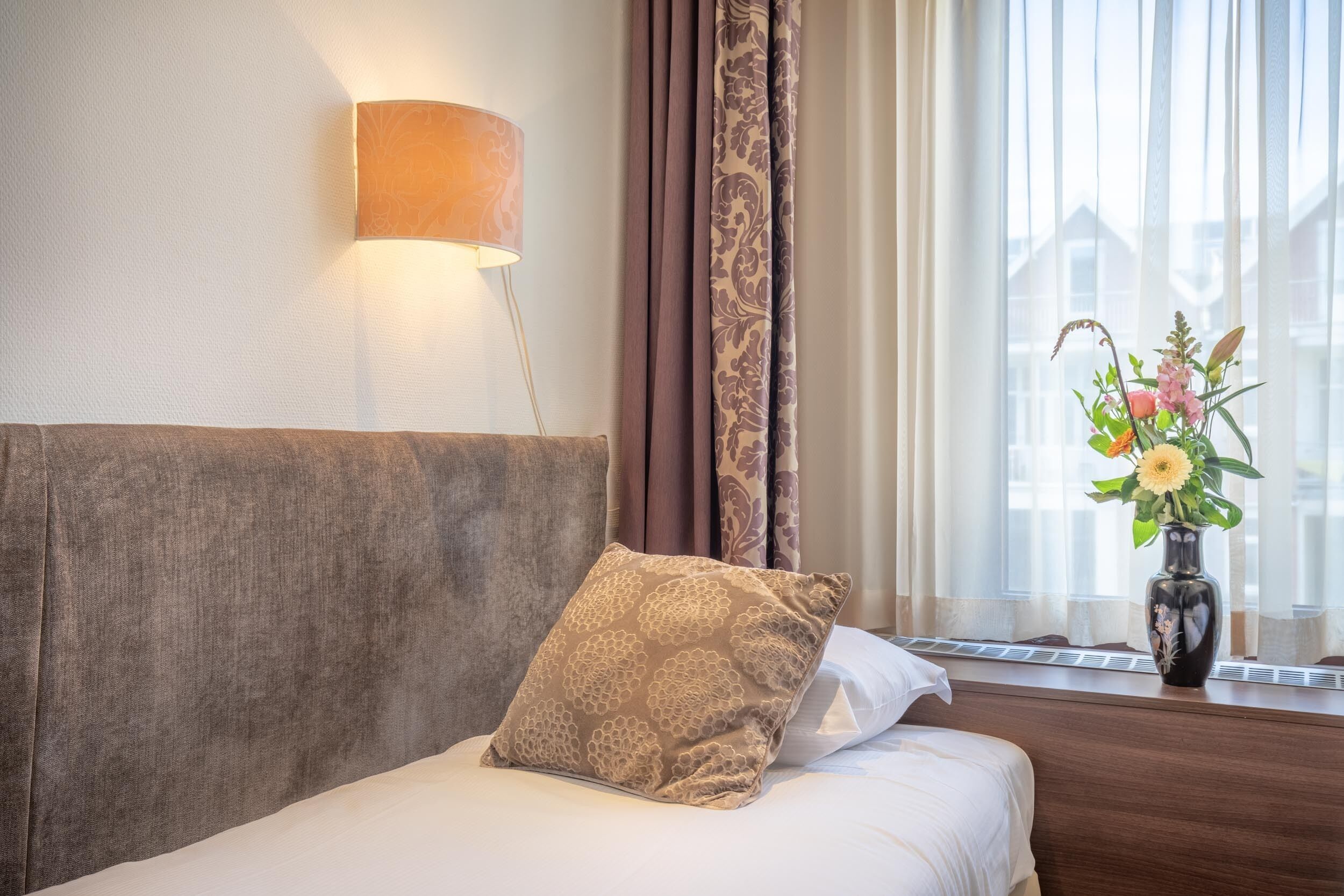 A hotel room with a bed, a wall-mounted lamp, a window with curtains, and a vase with flowers on the windowsill.