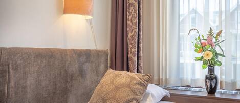 A hotel room with a bed, a wall-mounted lamp, a window with curtains, and a vase with flowers on the windowsill.