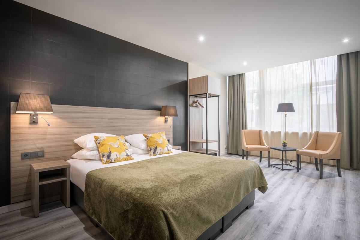 A modern hotel room with a large bed, bedside lamps, a small table, two chairs, and a wardrobe.