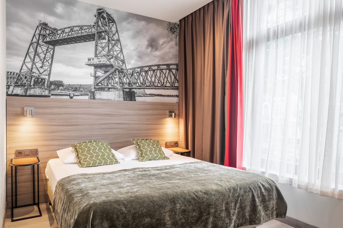 A hotel room with a large bed, two bedside tables, a headboard with a mural of a bridge, and a window with sheer curtains.