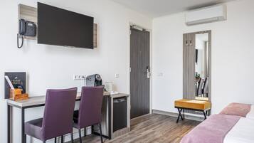 Een hotelkamer met een bureau, twee stoelen, een klein keukenhoekje, een bed, een spiegel en een aan de muur bevestigde televisie.