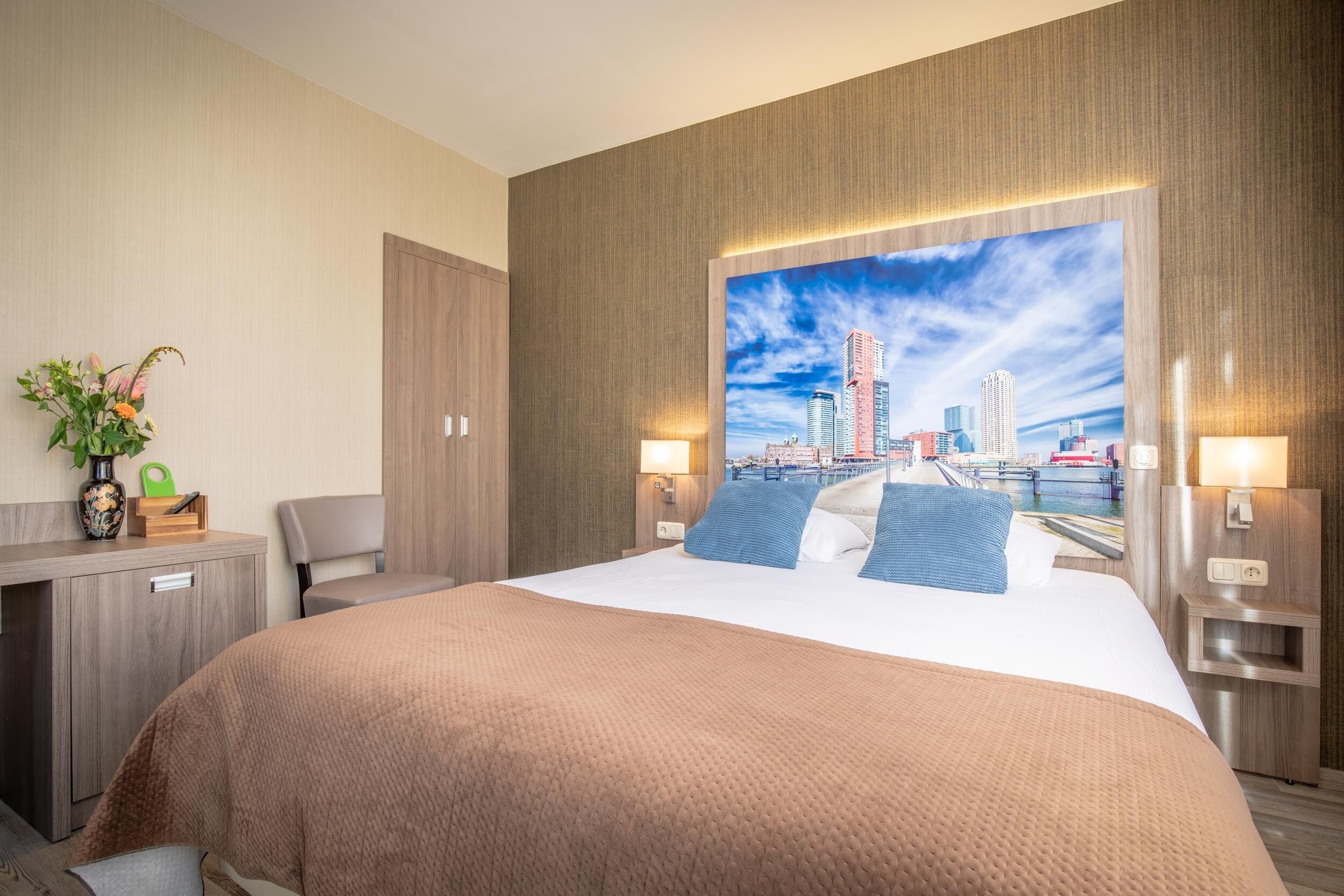 A hotel room with a large bed, a desk, a chair, a nightstand, and a cityscape view.
