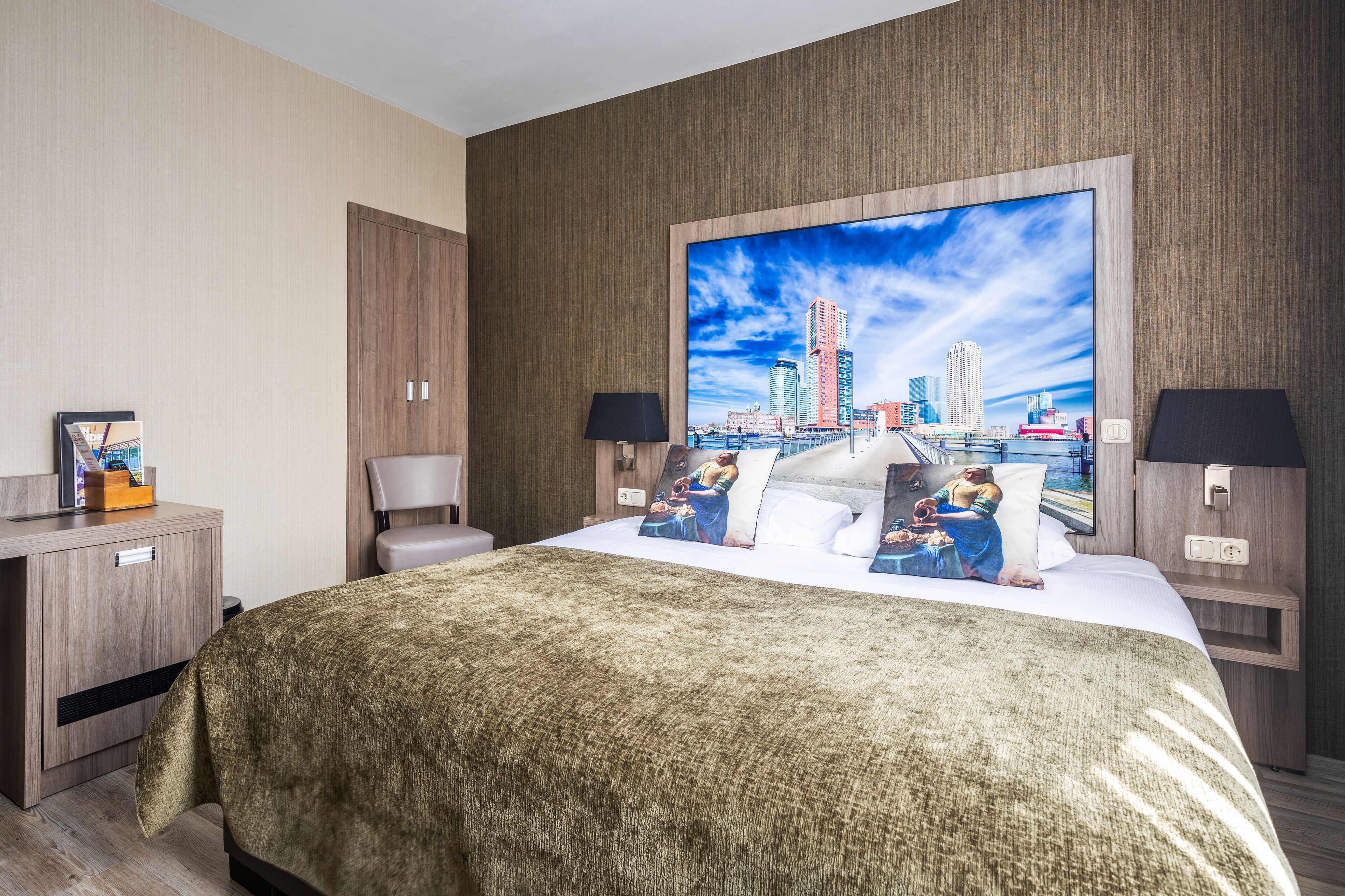 A hotel room with a large bed, a desk with a lamp, and a cityscape painting on the wall.