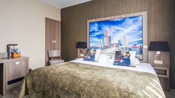 A hotel room with a large bed, a desk with a lamp, and a cityscape painting on the wall.