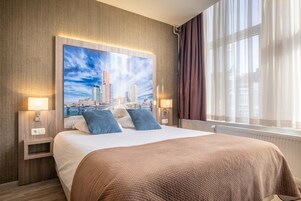Habitación de hotel con una cama grande, un cuadro de un paisaje urbano en la pared, dos lámparas de noche y una ventana con cortinas.