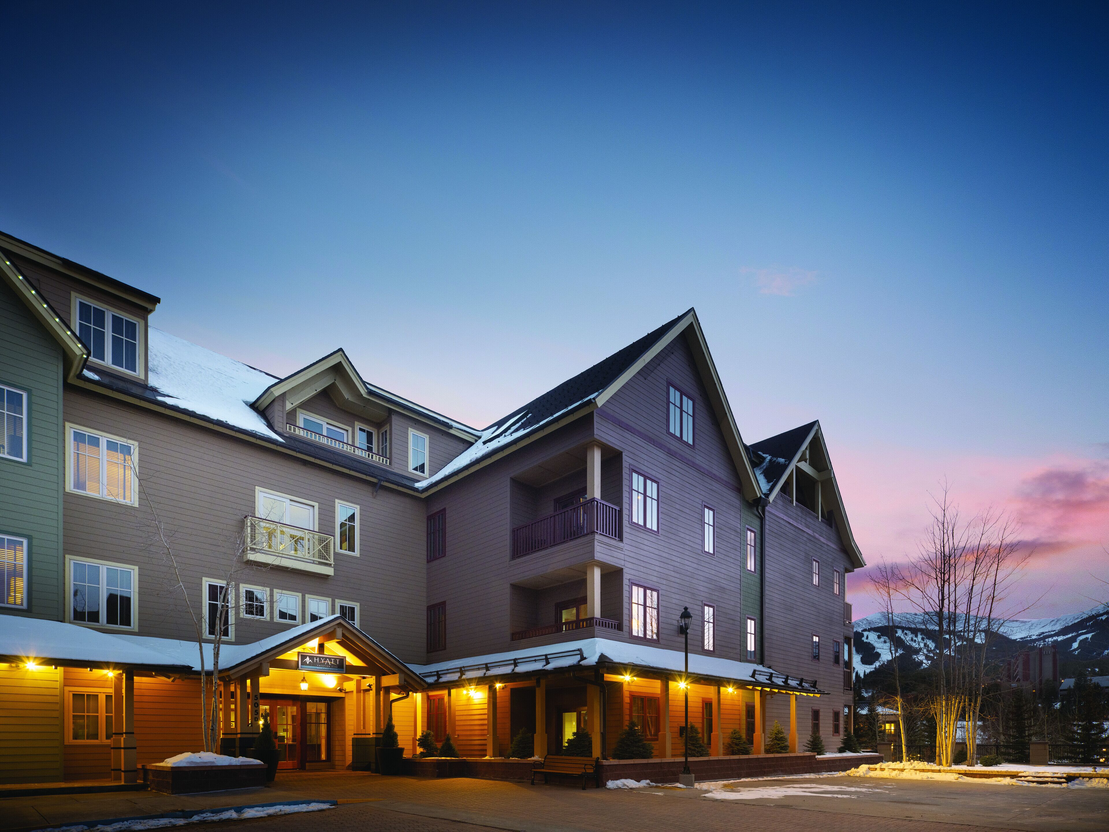 Ett hotell med flera våningar, ett väl synligt skylt, en överbyggd entré och utsikt över berg i bakgrunden.
