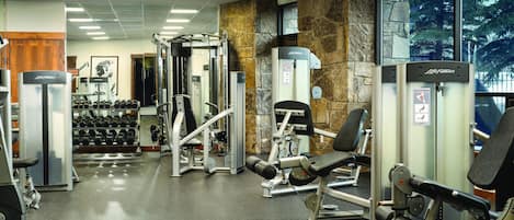 Une salle de sport équipée de divers appareils d’exercice, un mur orné d’un motif en pierre et une grande fenêtre donnant sur des arbres.