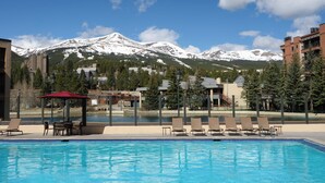 Una piscina con sillas de descanso y una sombrilla roja, rodeada de edificios y montañas nevadas.